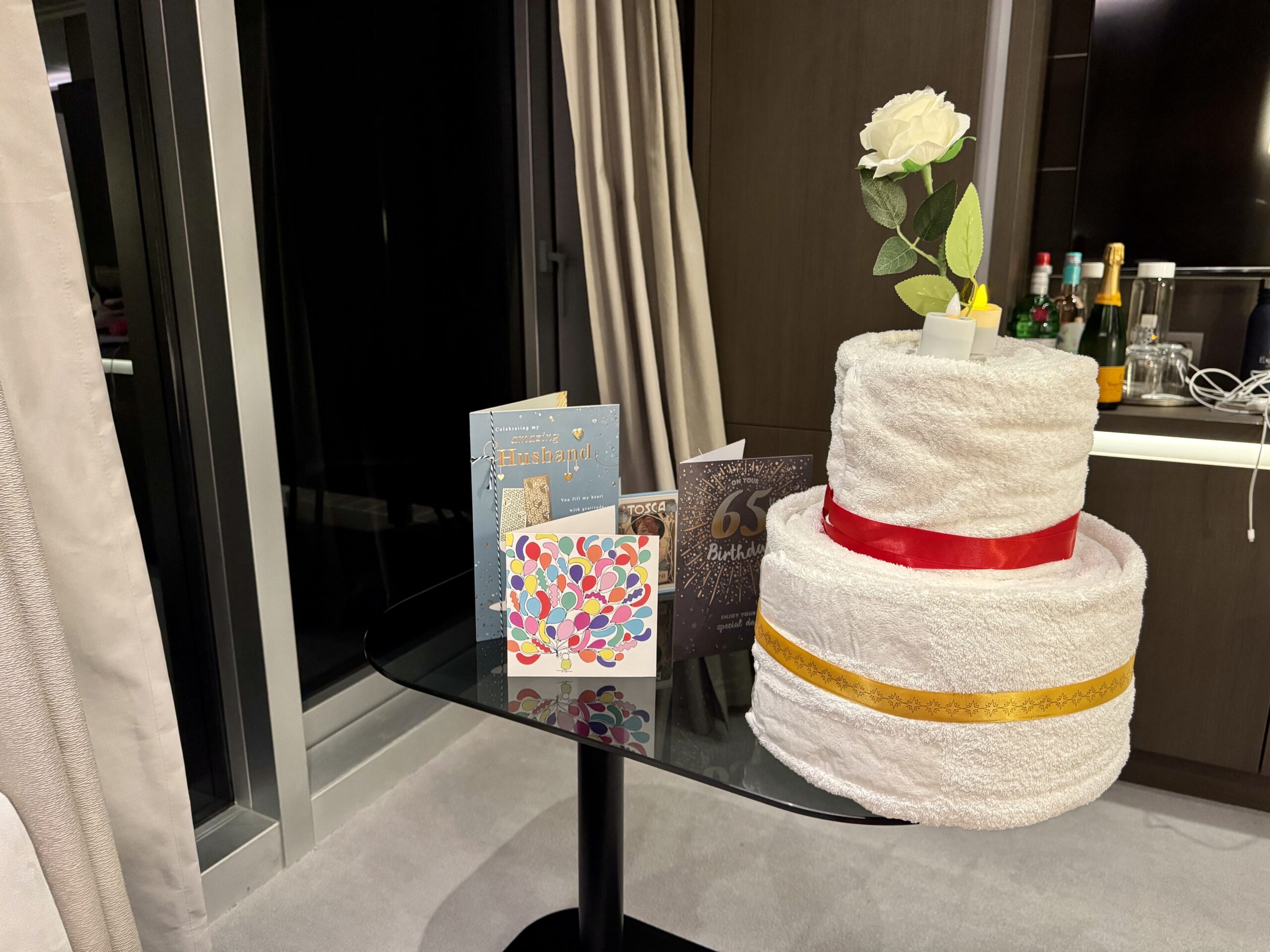 White towel cake with two tiers and a red ribbon on a glass table, with birthday cards nearby and a white rose stem in the background.