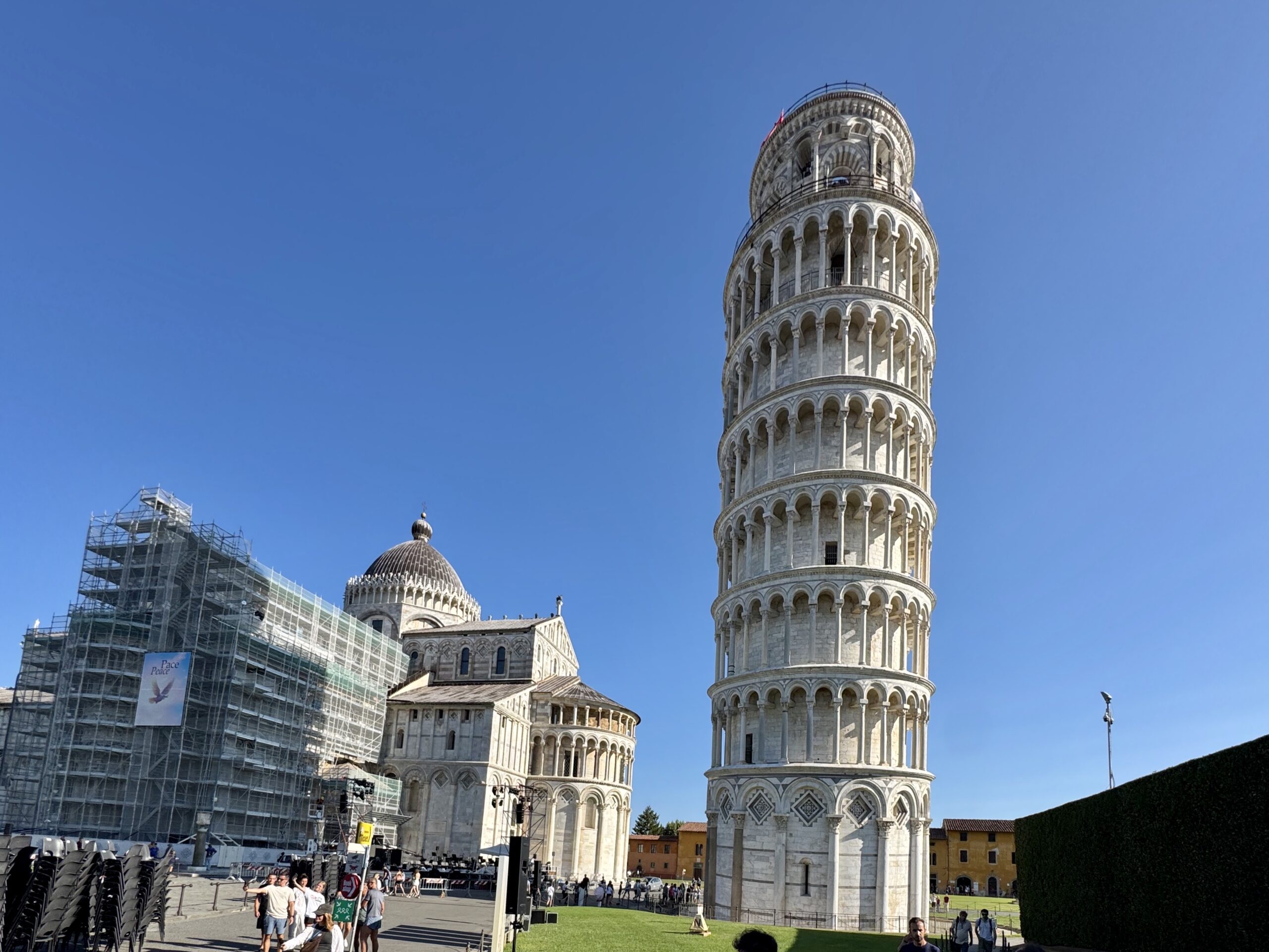 Leaning Tower of Pisa against a clear blue sky with scaffolding on the left and visitors in the foreground.