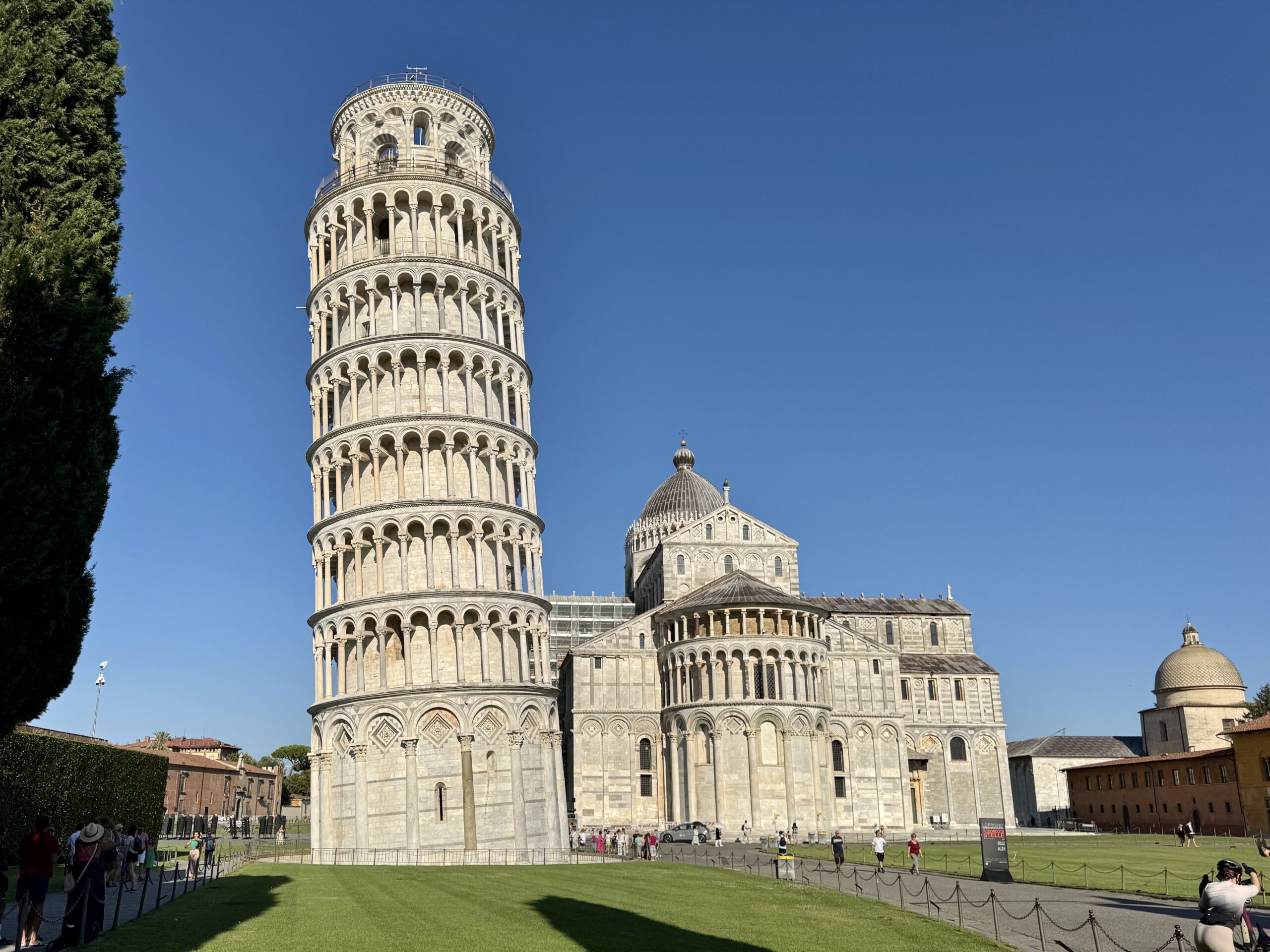 Leaning Tower of Pisa beside the cathedral complex against a blue sky, with visitors walking on the lawn in the foreground.