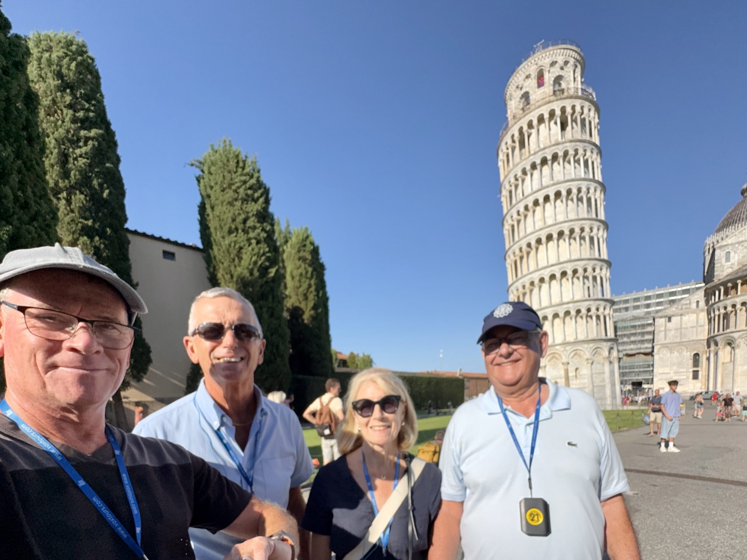 Group of tourists posing for a selfie with the Leaning Tower of Pisa in bright sun behind them; everyone wears lanyards and smiles.