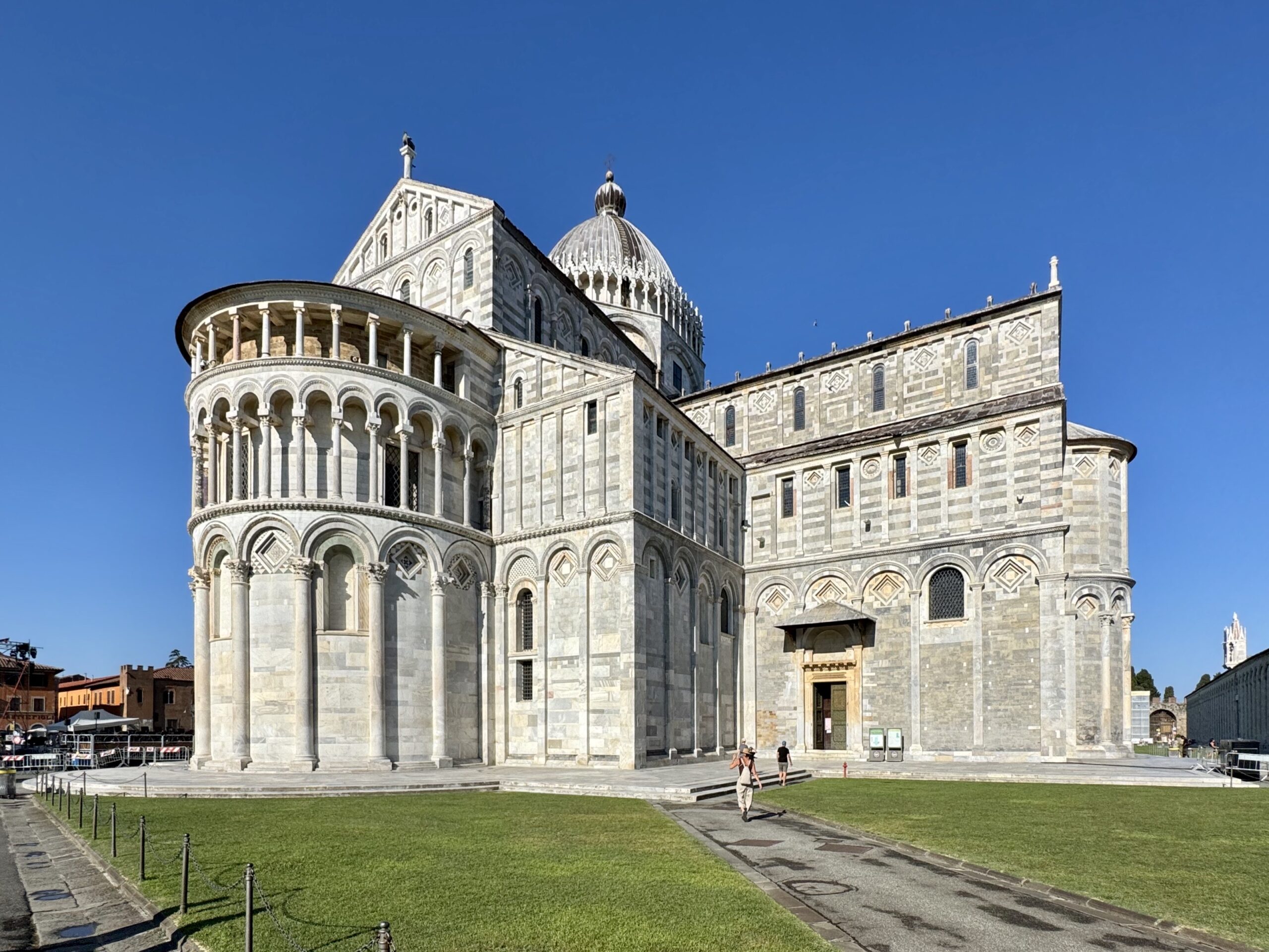 Stone cathedral with rounded arches and a large striped dome, set against a clear blue sky; foreground lawn and stone walkway with visitors nearby.