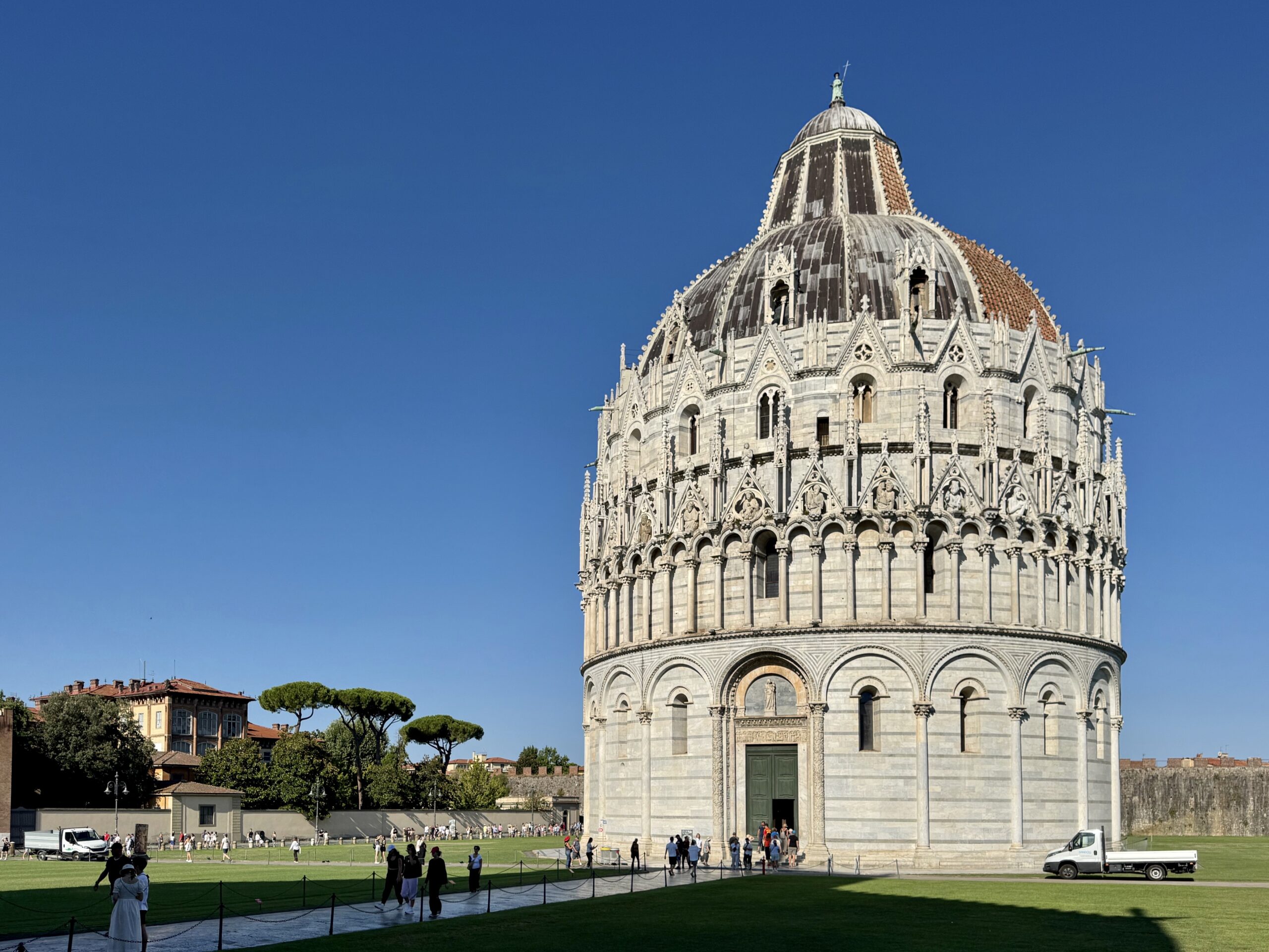 Dome of Pisa Cathedral with ornate stonework, visitors on the lawn in Piazza dei Miracoli under a clear blue sky.