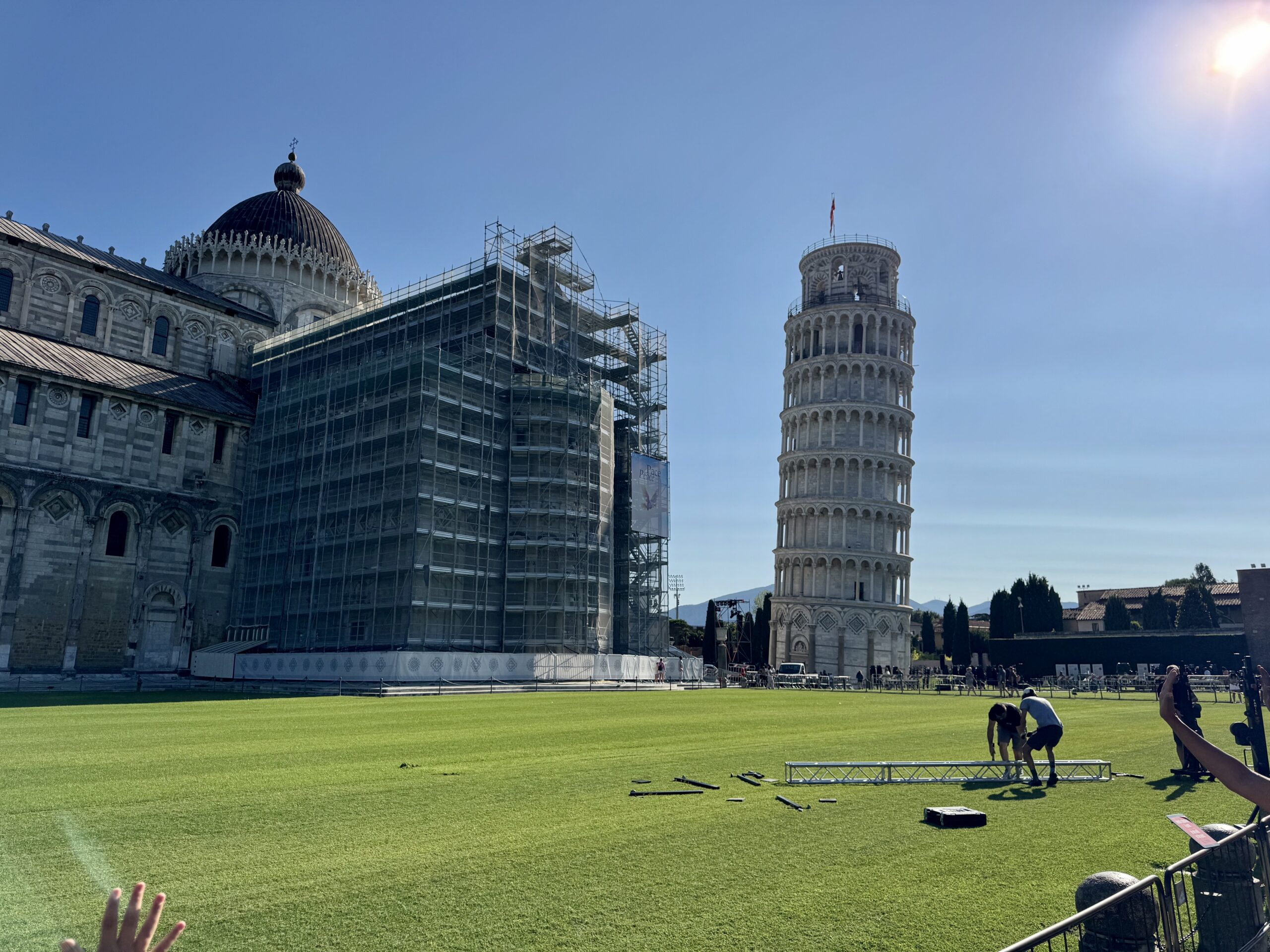 Leaning Tower of Pisa beside a stone building with scaffolding on a sunny day, people on a green lawn preparing equipment.