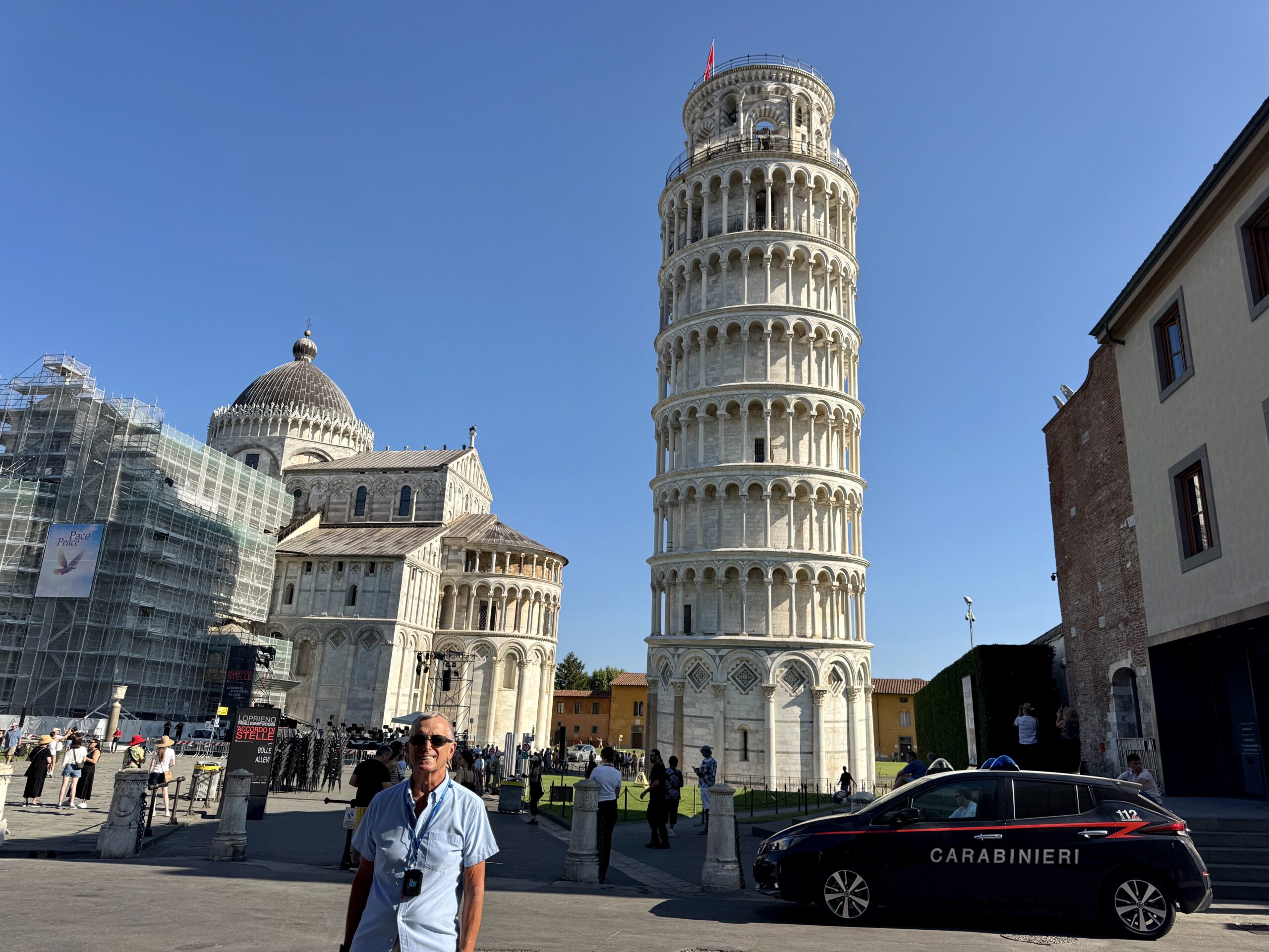 Tourists near the Leaning Tower of Pisa on a sunny day with a Carabinieri car in the foreground