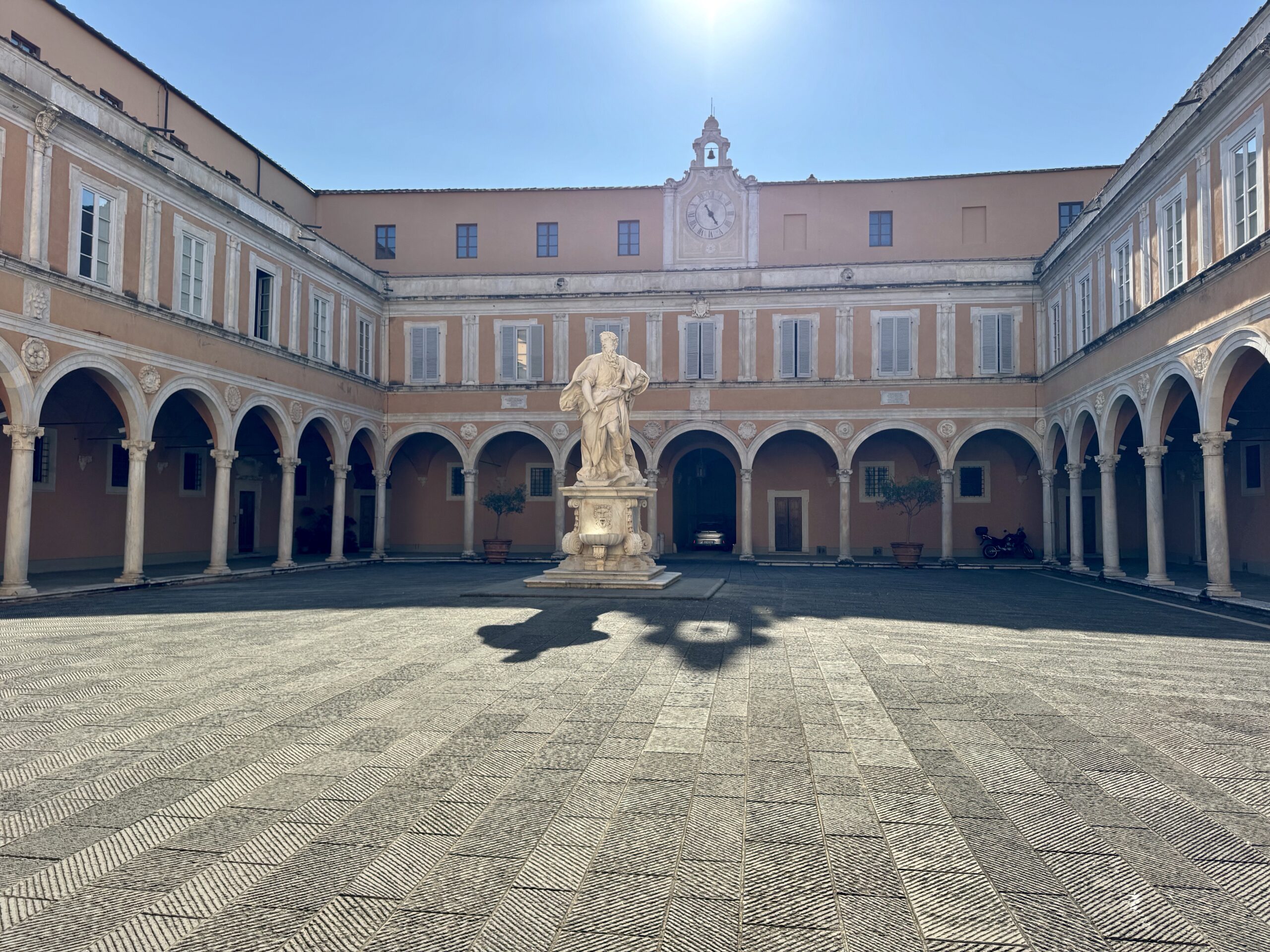 Courtyard with pale peach walls, arched colonnade, and a central statue fountain beneath a clock tower.