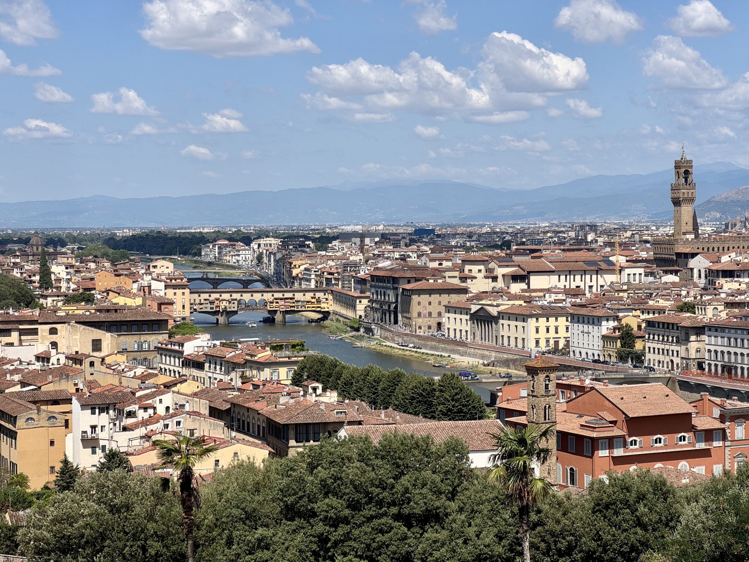 Panoramic view of a historic Italian city along a river, with orange rooftops, arched bridges, and a tall brick tower under a blue sky.