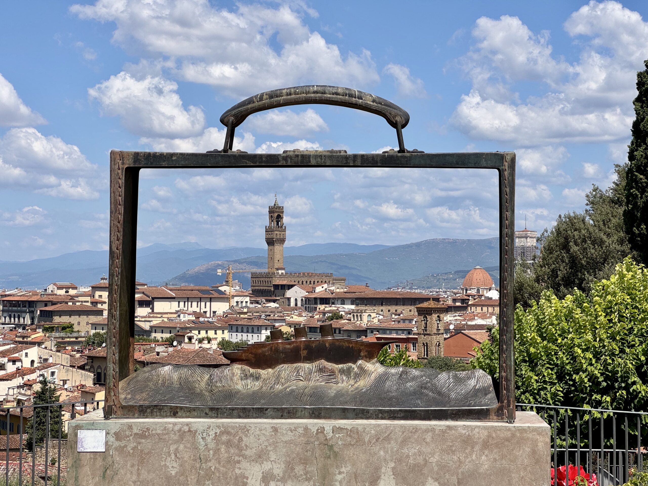 Rusty metal frame framing a cityscape with terracotta rooftops and a tall tower under a blue sky with clouds.