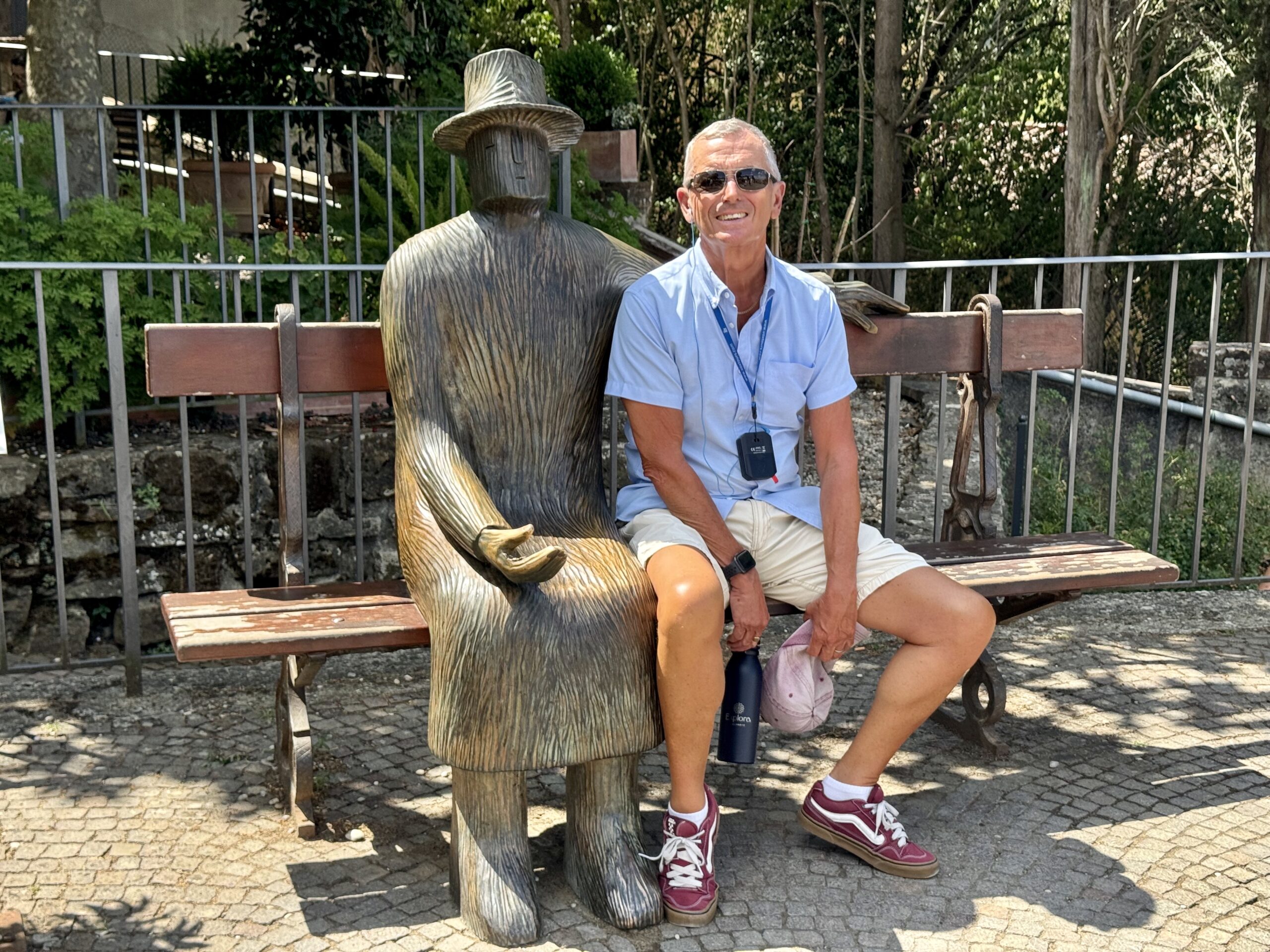 Man in sunglasses sits on a bench beside a tall carved wooden statue wearing a hat in a sunny outdoor area behind a railing.