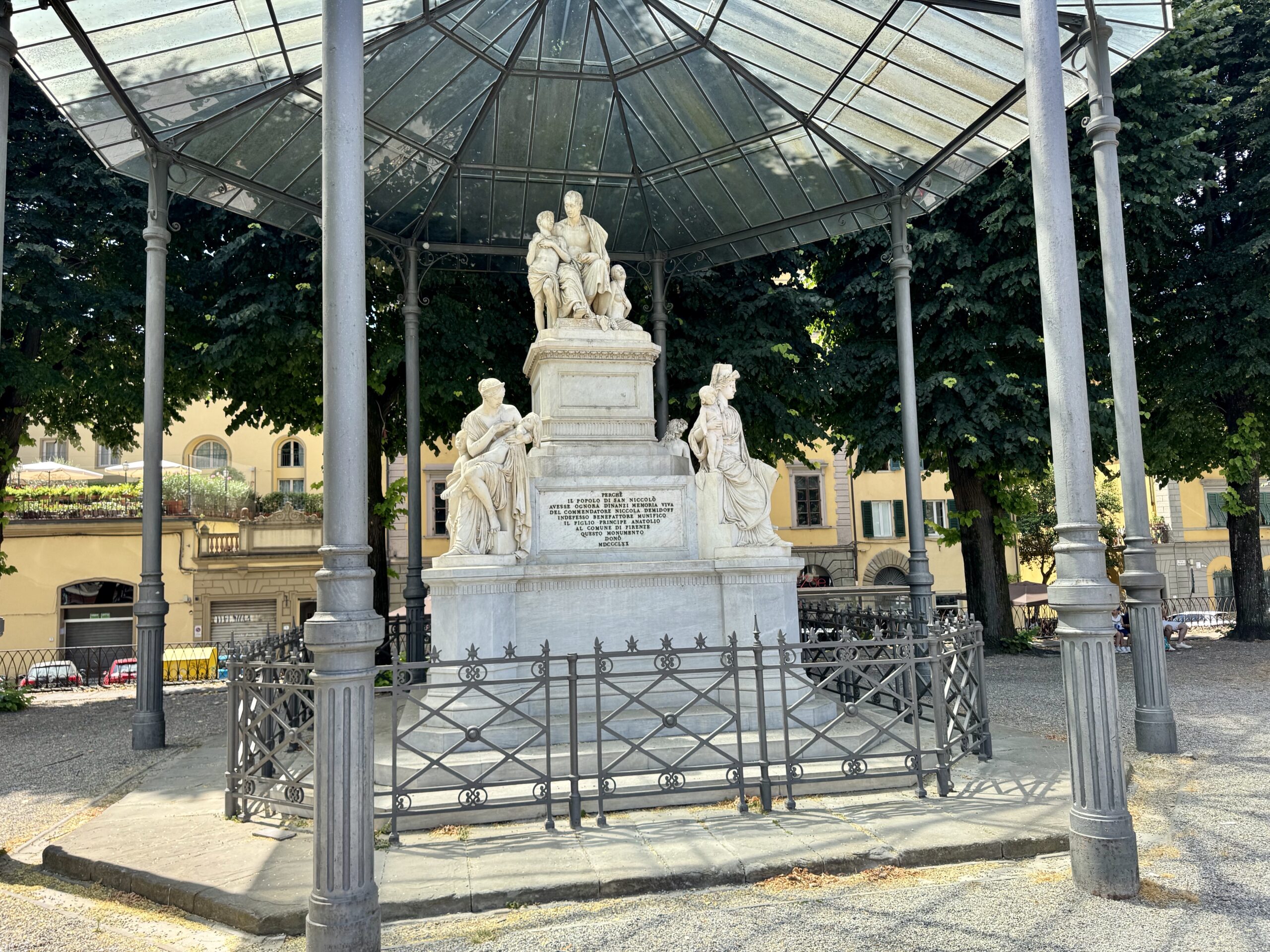Marble statue group on a pedestal under a glass and iron pavilion in a square, with green trees and yellow buildings in the background.