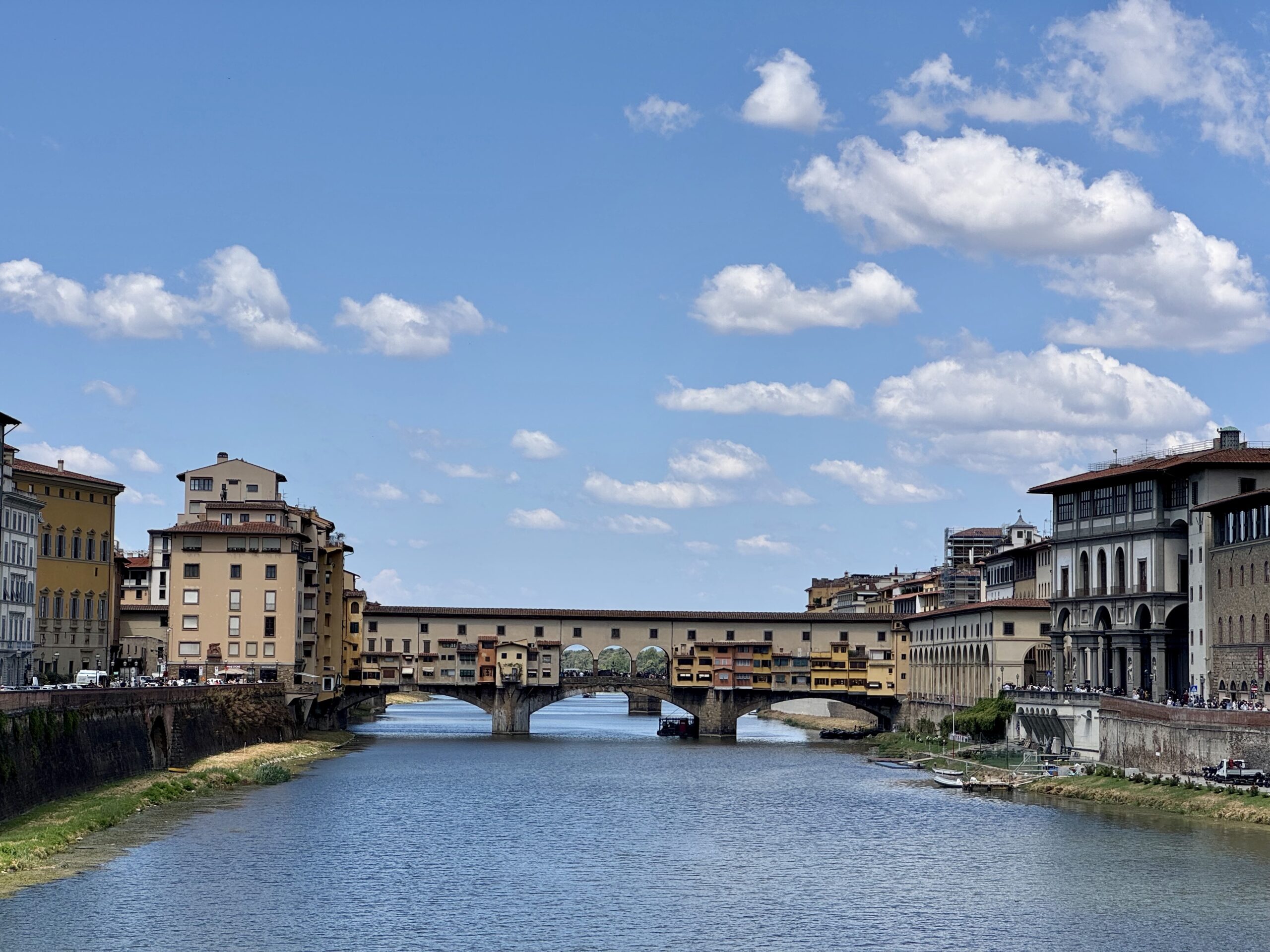 Riverside cityscape with a stone arched bridge over a calm river and pastel-colored buildings on both sides under a bright blue sky with scattered clouds.