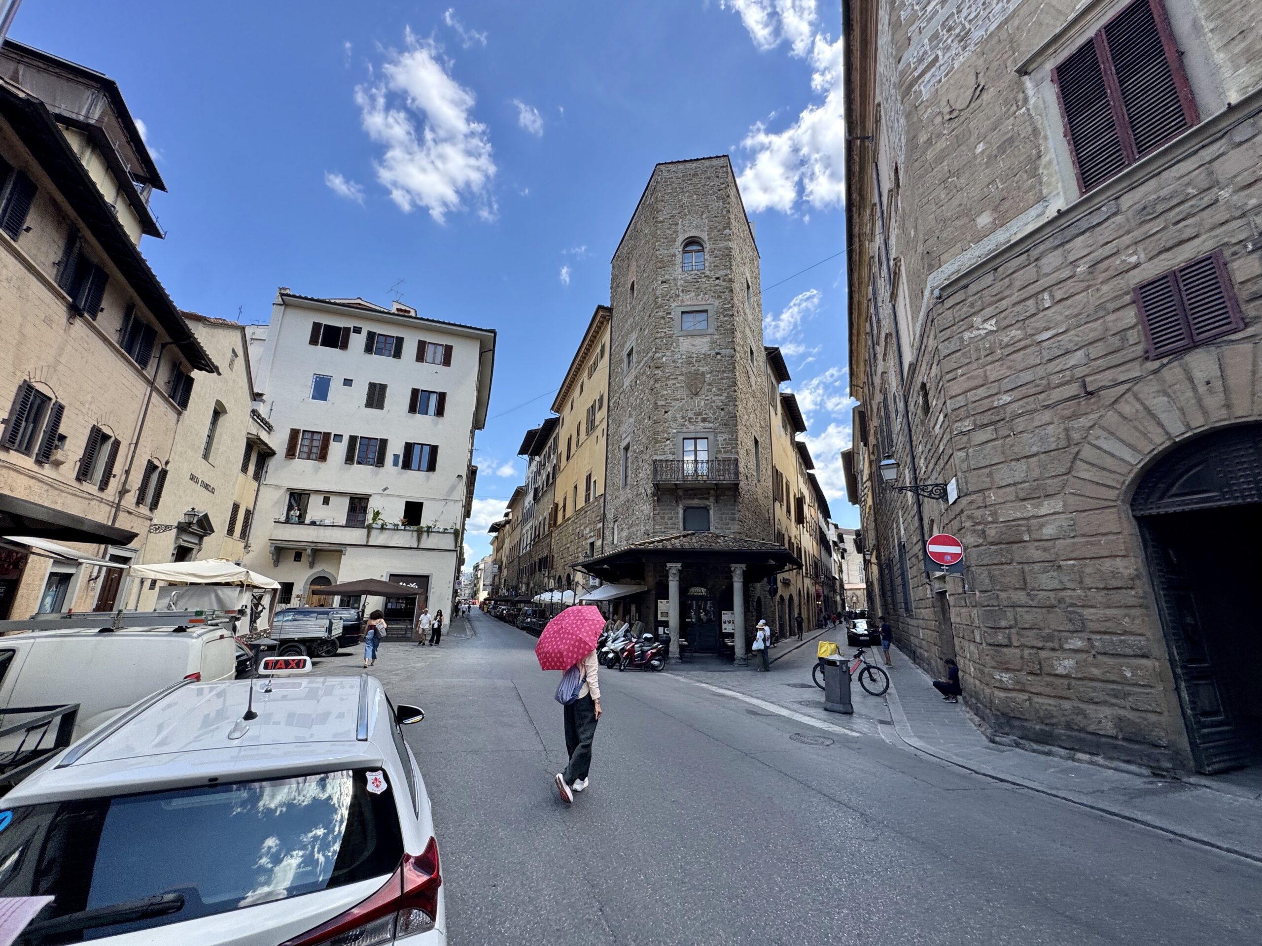 Narrow cobblestone street lined with historic stone buildings under a bright blue sky; pedestrians walk, some with umbrellas, and parked cars along the curb.
