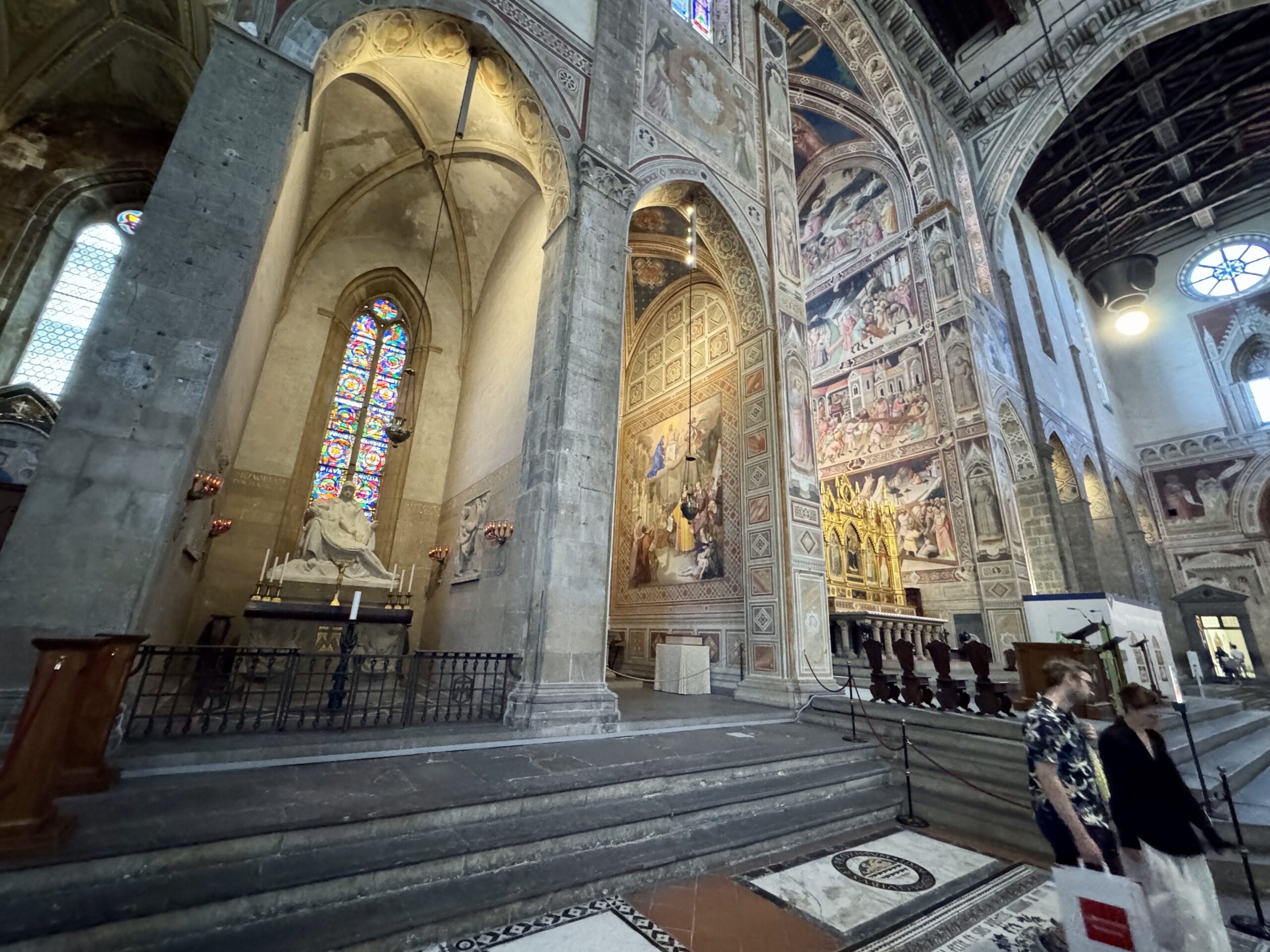 Interior of a historic cathedral with ornate frescoed walls, stone arches, and stained-glass window panels along the aisle outside pews and railings.