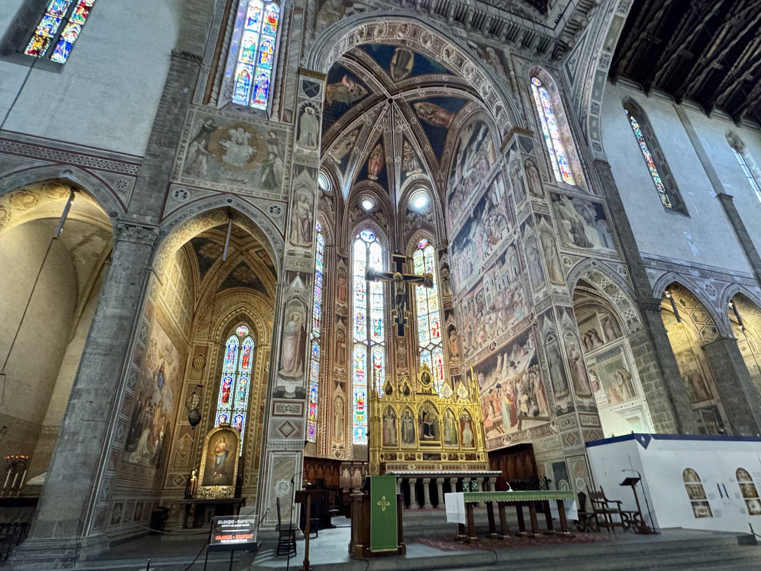Interior of a grand Gothic church with ornate arches, frescoed walls, and tall stained-glass windows surrounding a lit altar.