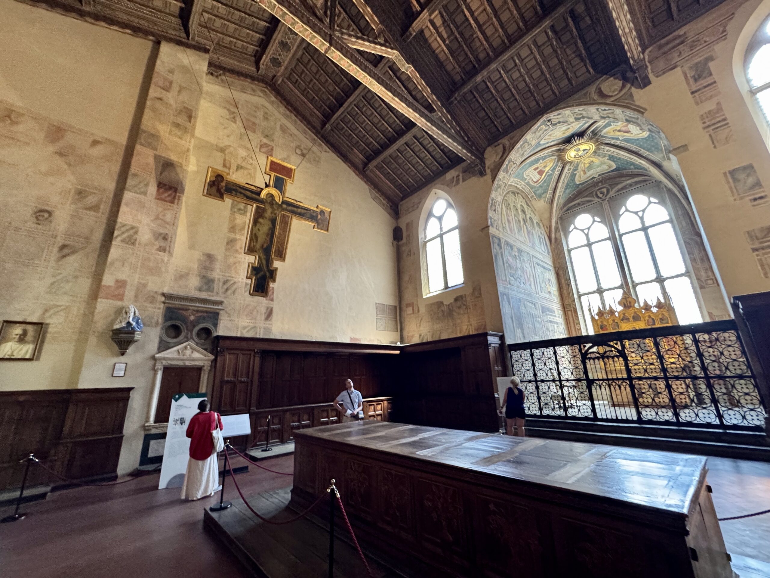 Interior of a historic church: hanging crucifix, dark wooden pews, stone walls, and tall arched stained-glass windows.