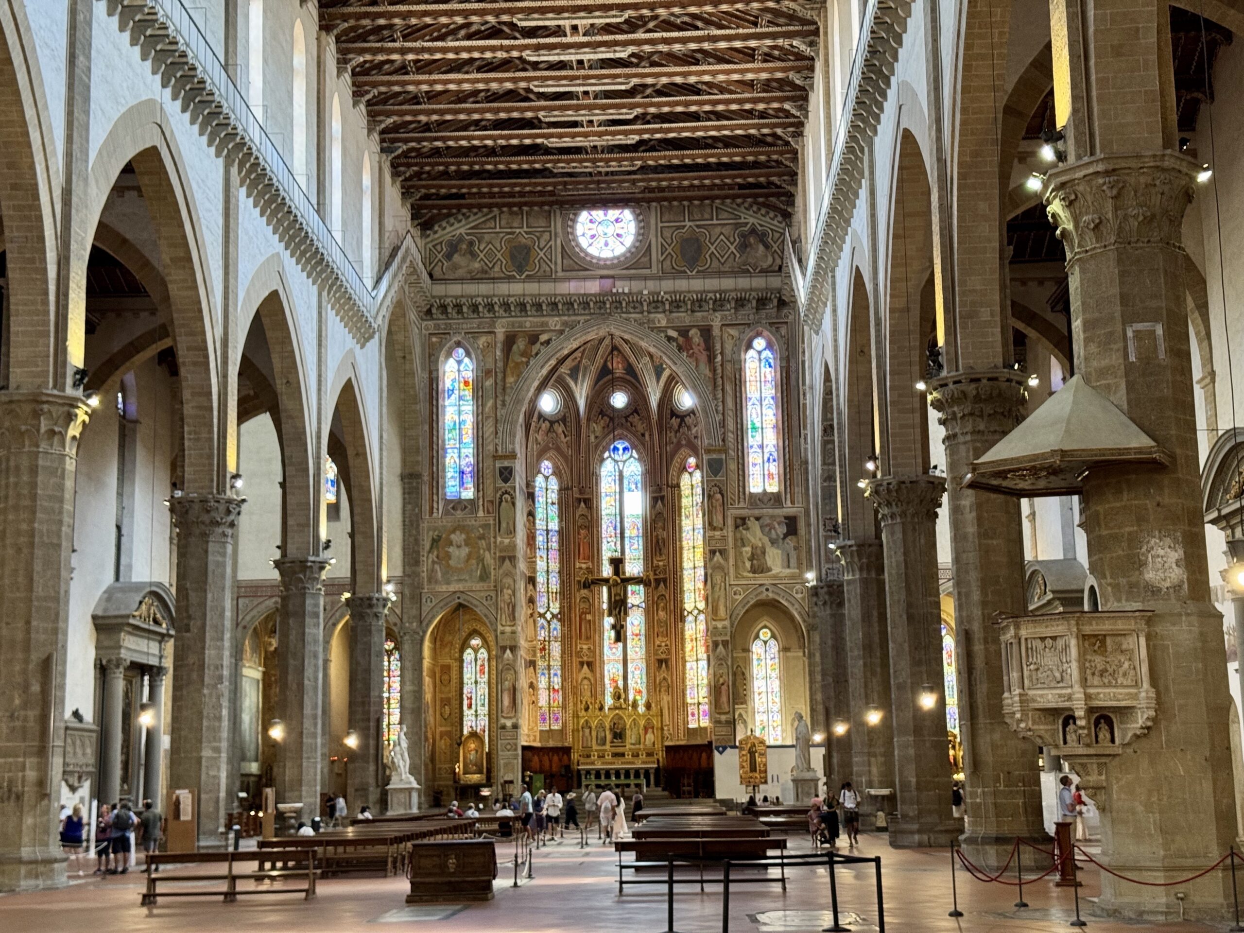 Interior of a grand cathedral nave with tall arches, ornate columns, and a wooden vaulted ceiling; visitors seated along pews.