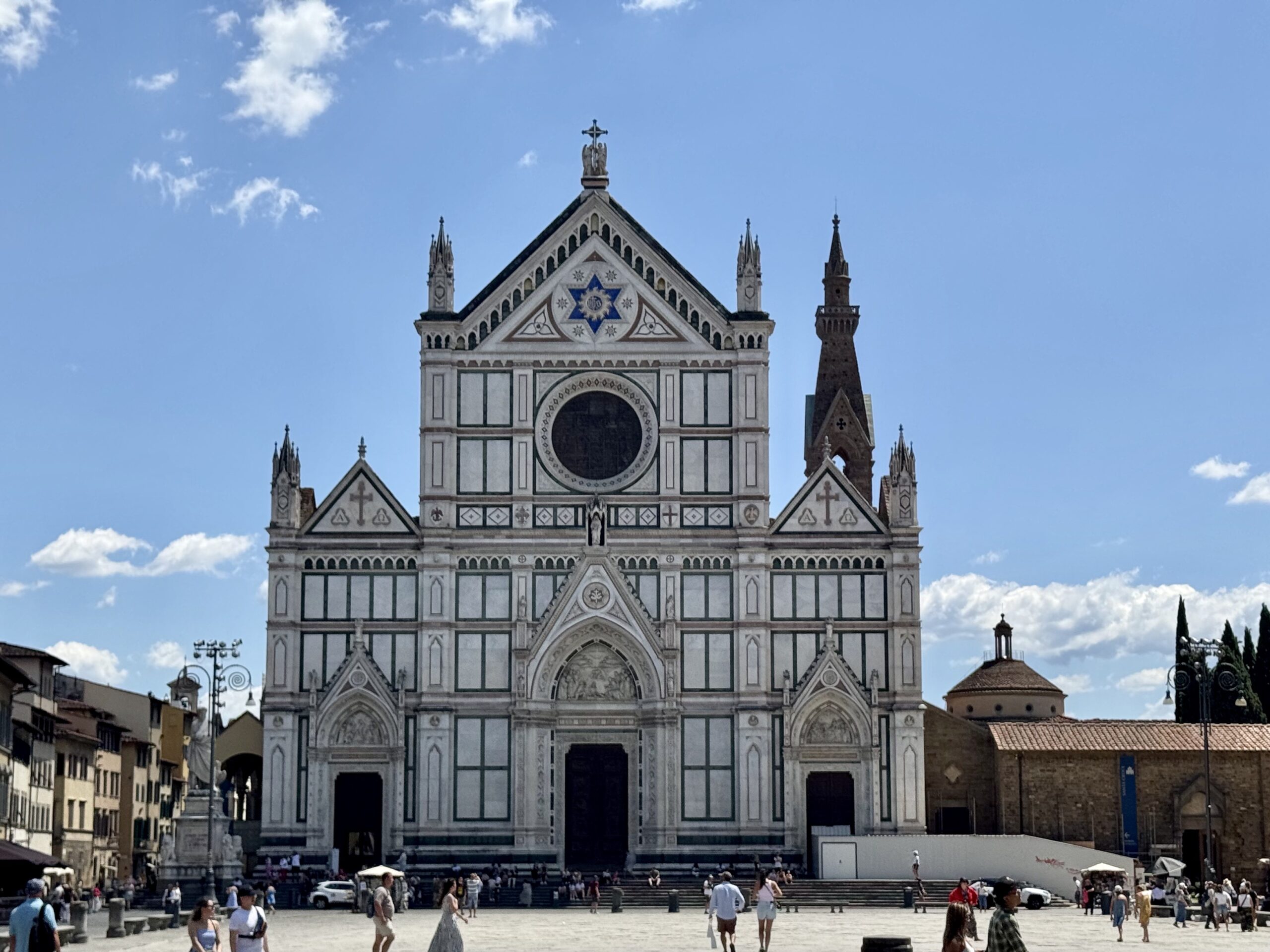 Front façade of the cathedral with white and green marble, a large circular window, and decorative arches in a busy square.