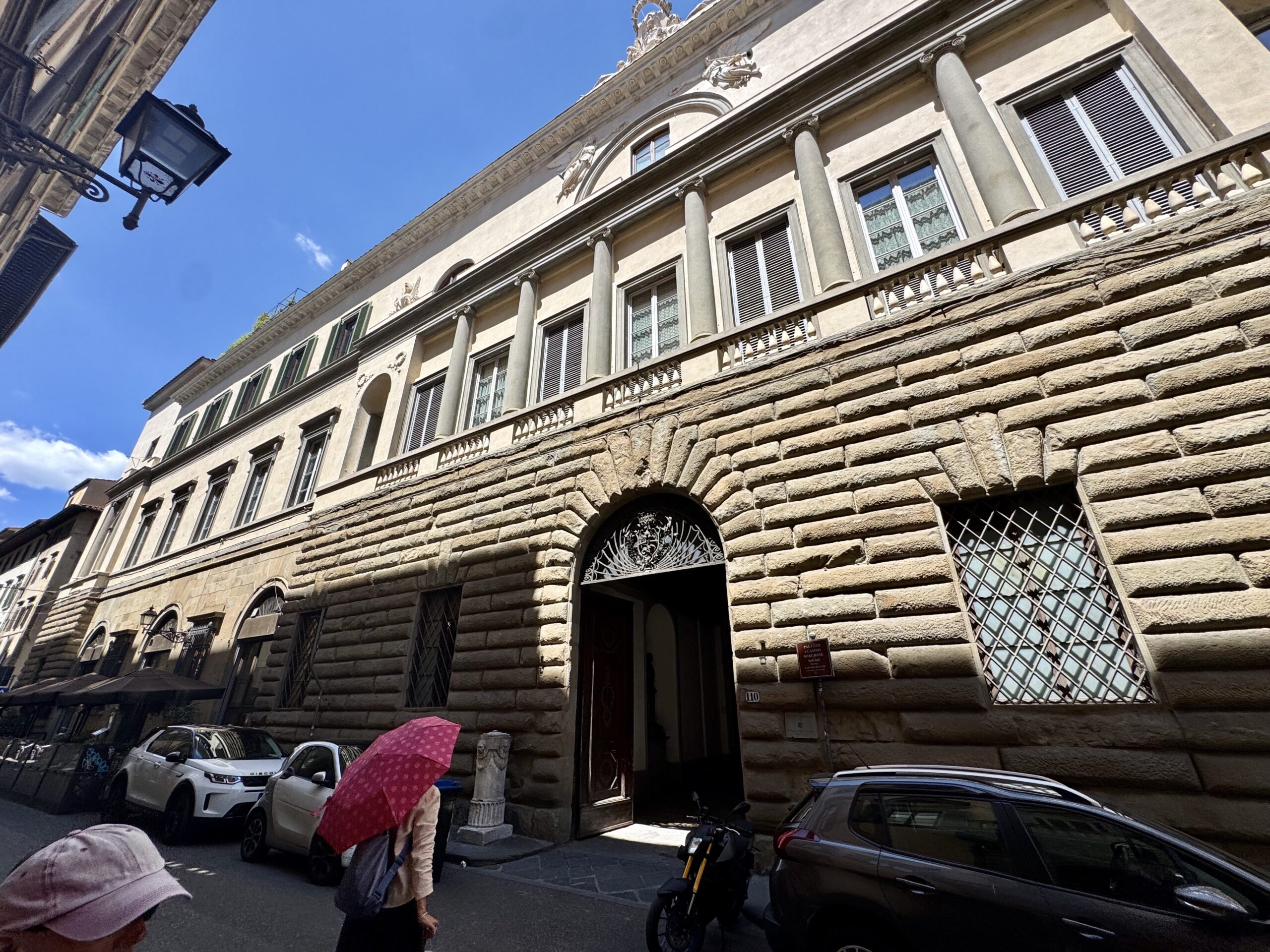 Stone neoclassical building with arched entrance, columns, and decorative façade under a bright blue sky.