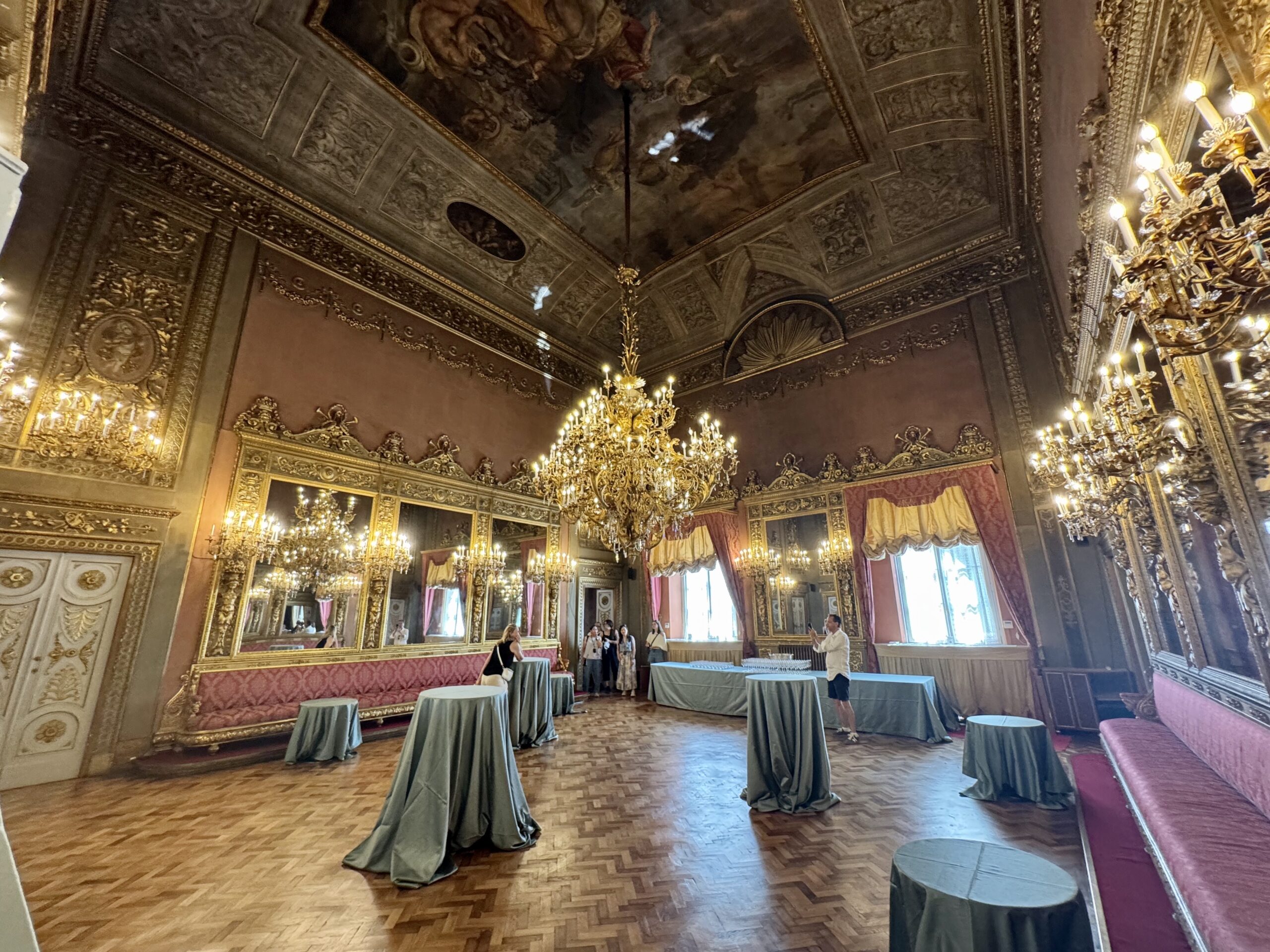 Opulent ballroom with gilded walls, ornate carvings, and crystal chandeliers; round cocktail tables covered in green cloths set up for an event.