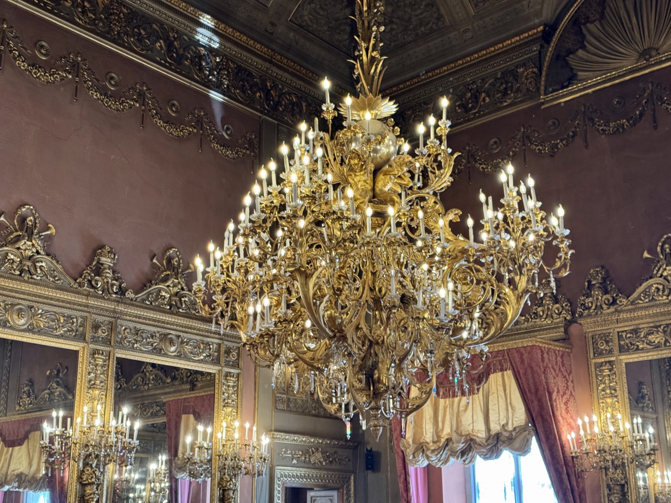 Opulent gold chandelier with numerous candles hangs over a richly carved, gilded interior room with burgundy walls and ornate trim.