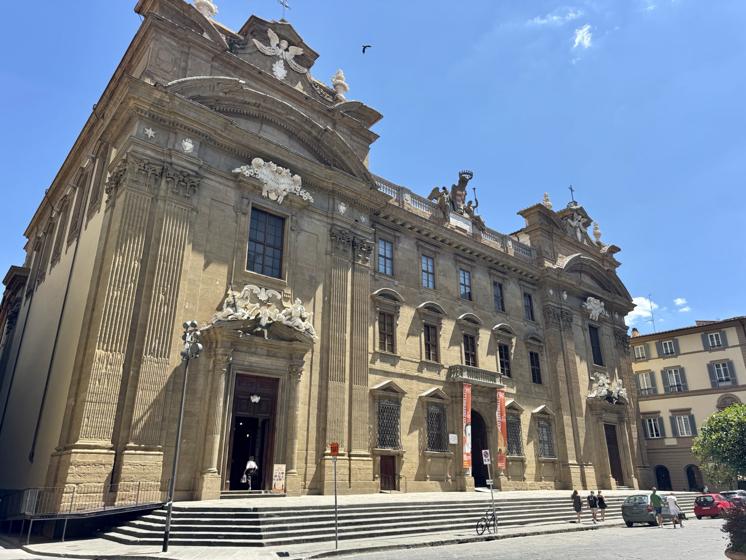 Facade of a historic stone building with ornate carvings, statues atop, and wide steps leading to the entrance under a clear blue sky.