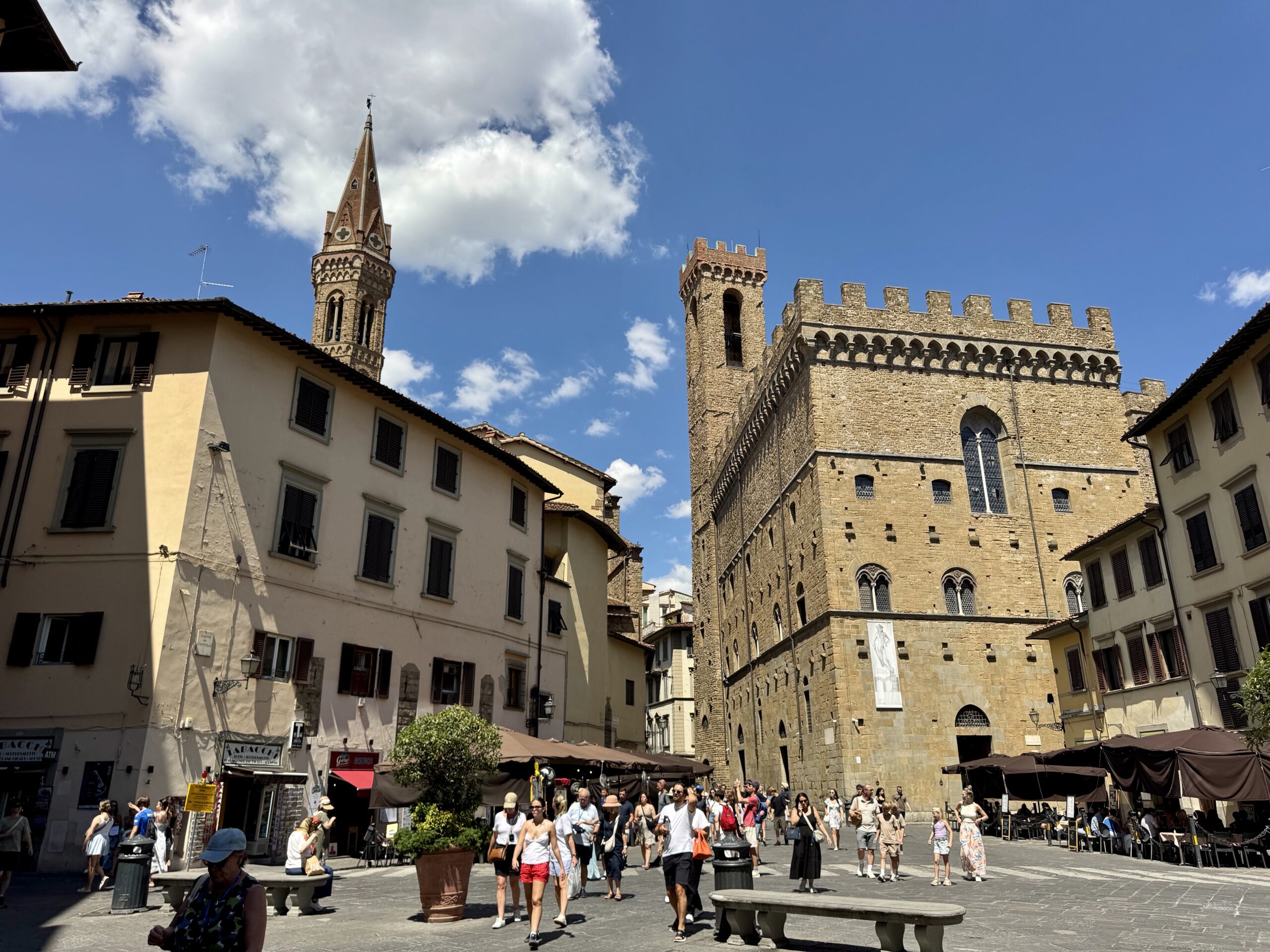 Historic town square with medieval stone buildings and a tall church spire under a blue sky; people walk and outdoor cafes line the street.