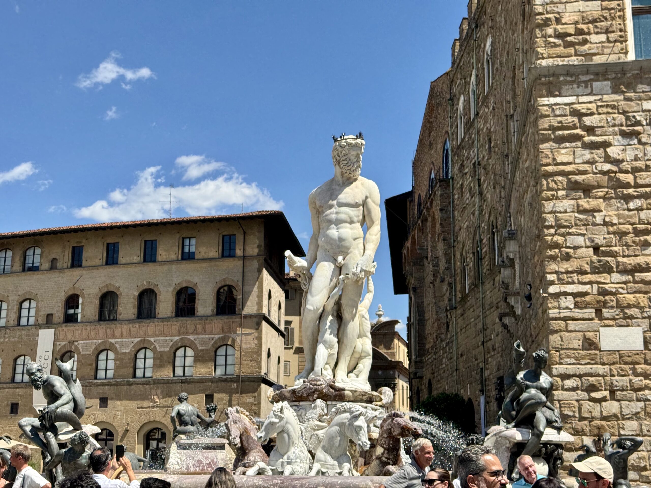 Central marble statue rising from a historic fountain, surrounded by other sculptures in a bustling square under a blue sky.