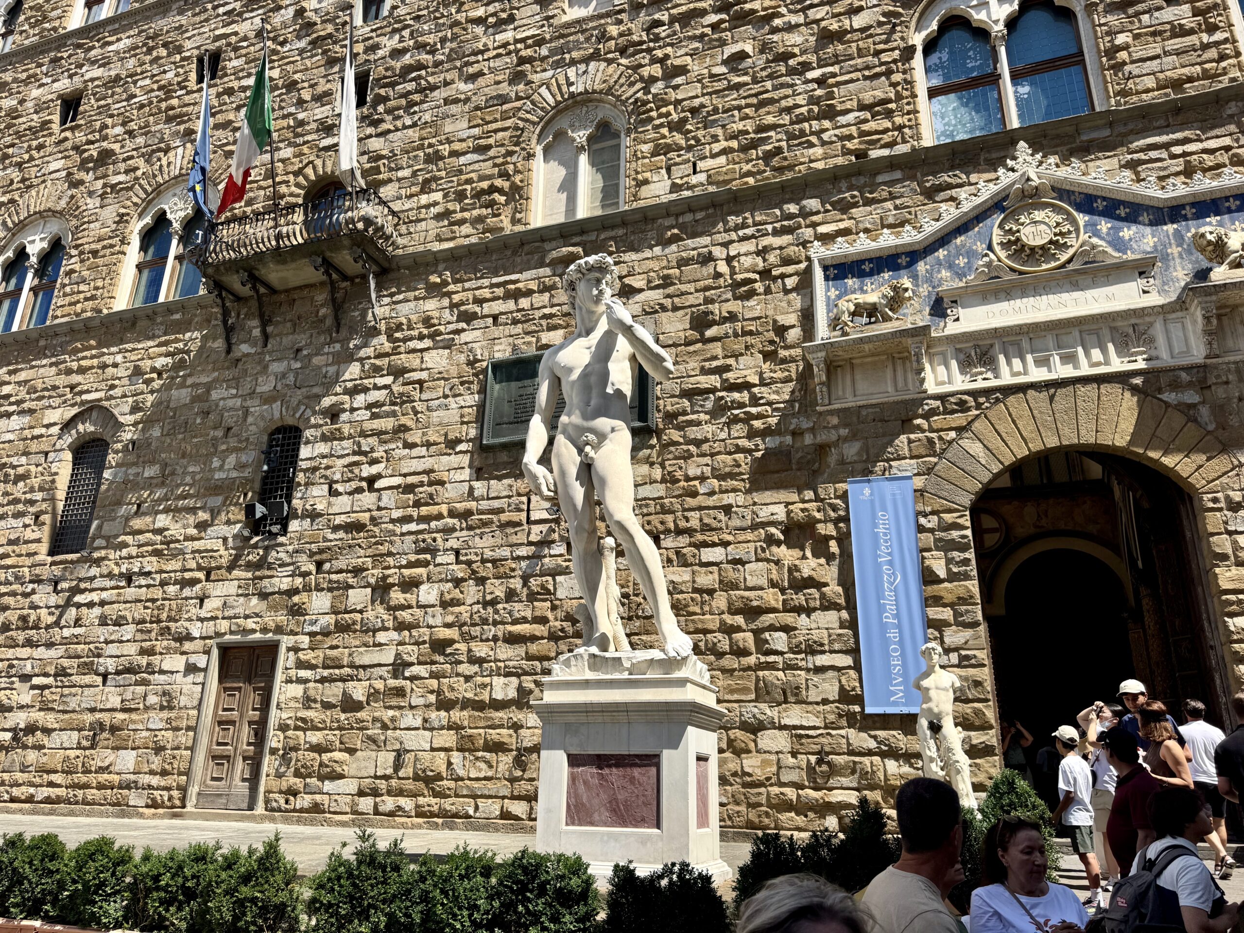Stone historic building with flags on a balcony, a prominent marble statue in the foreground, and visitors nearby.