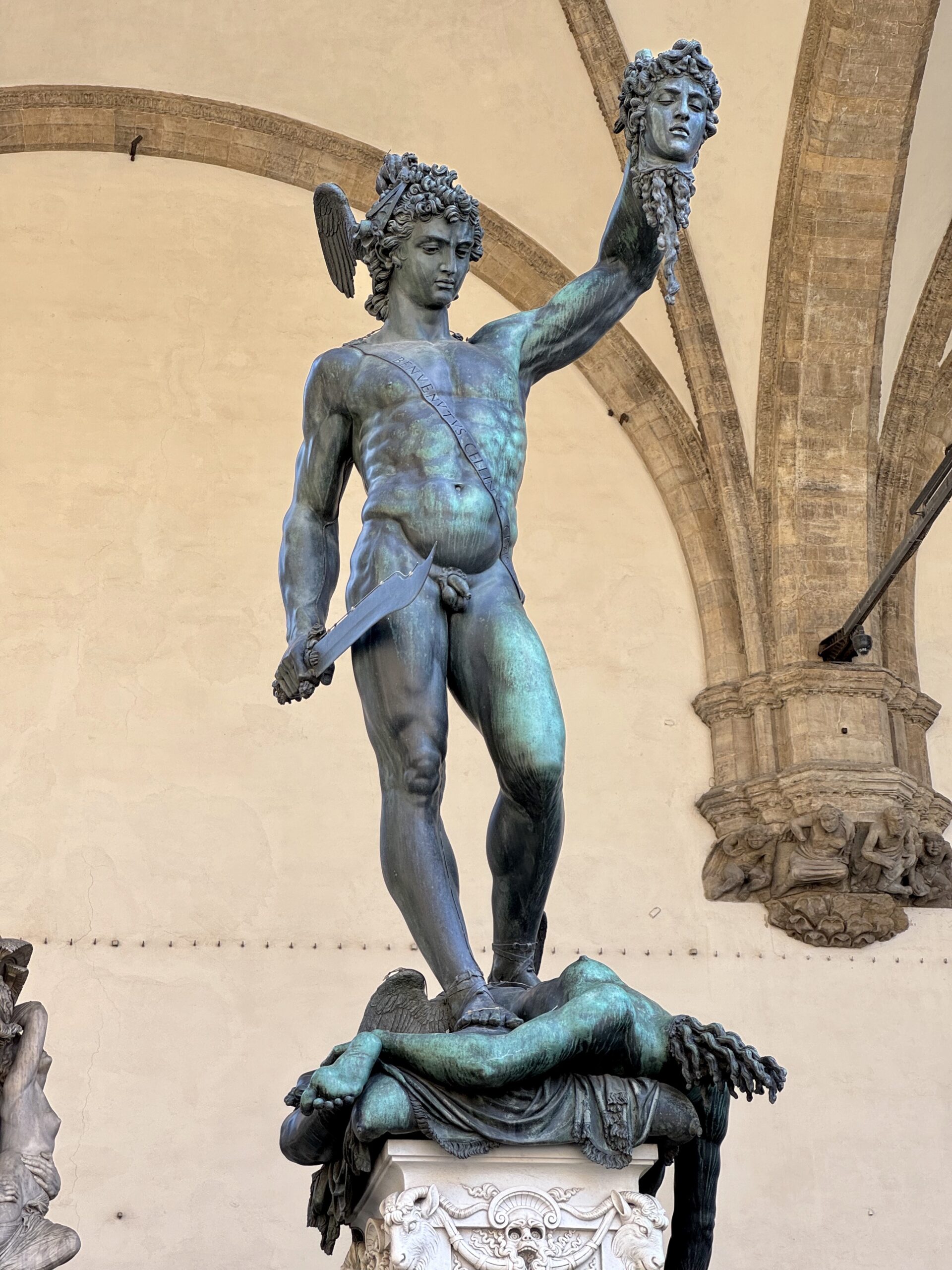 Bronze statue of Perseus holding Medusa's severed head aloft, standing on a carved pedestal in a pale courtyard.
