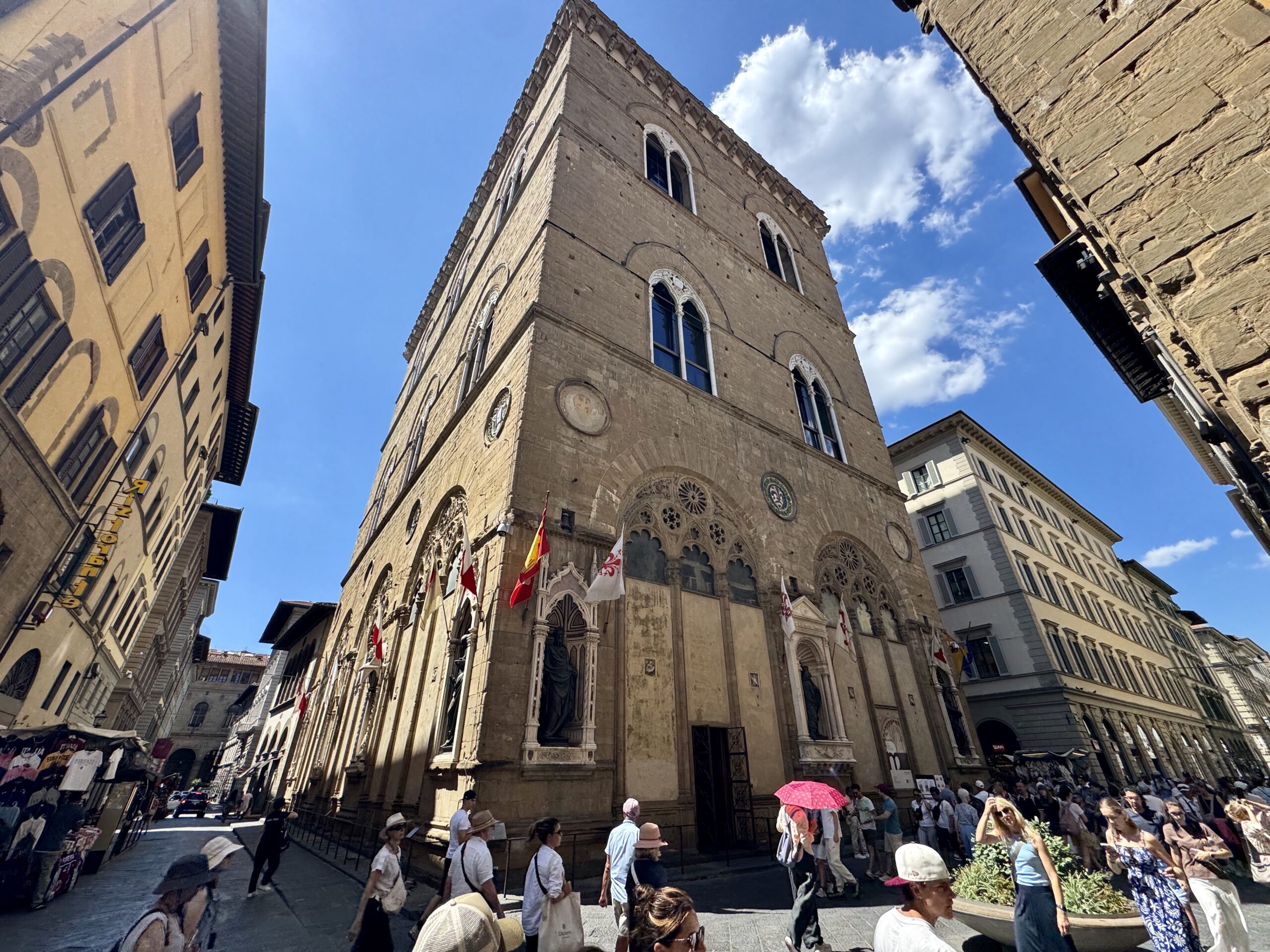 Historic stone church with arched windows on a sunny plaza, flags hanging along the facade as pedestrians pass by.