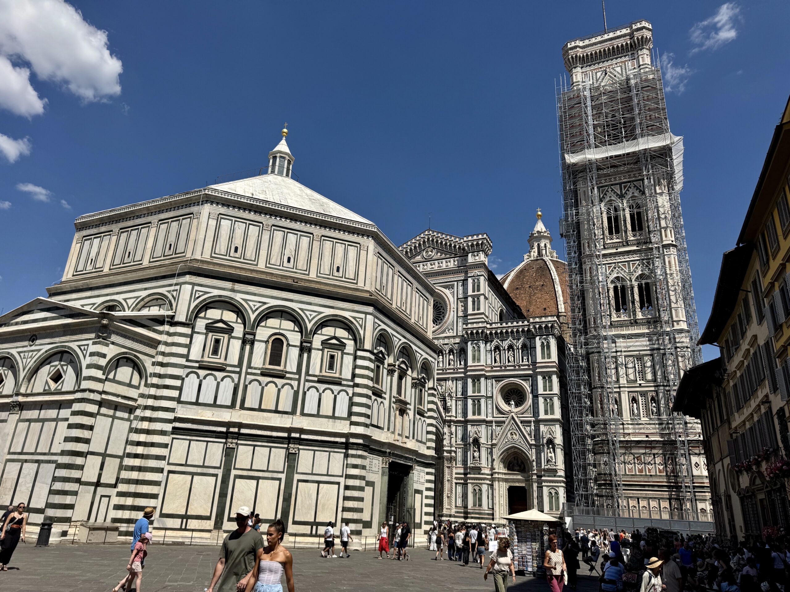 Facade of Florence Cathedral with green‑white marble stripes, red dome in the background, and a scaffolding‑wrapped bell tower; people in the square.