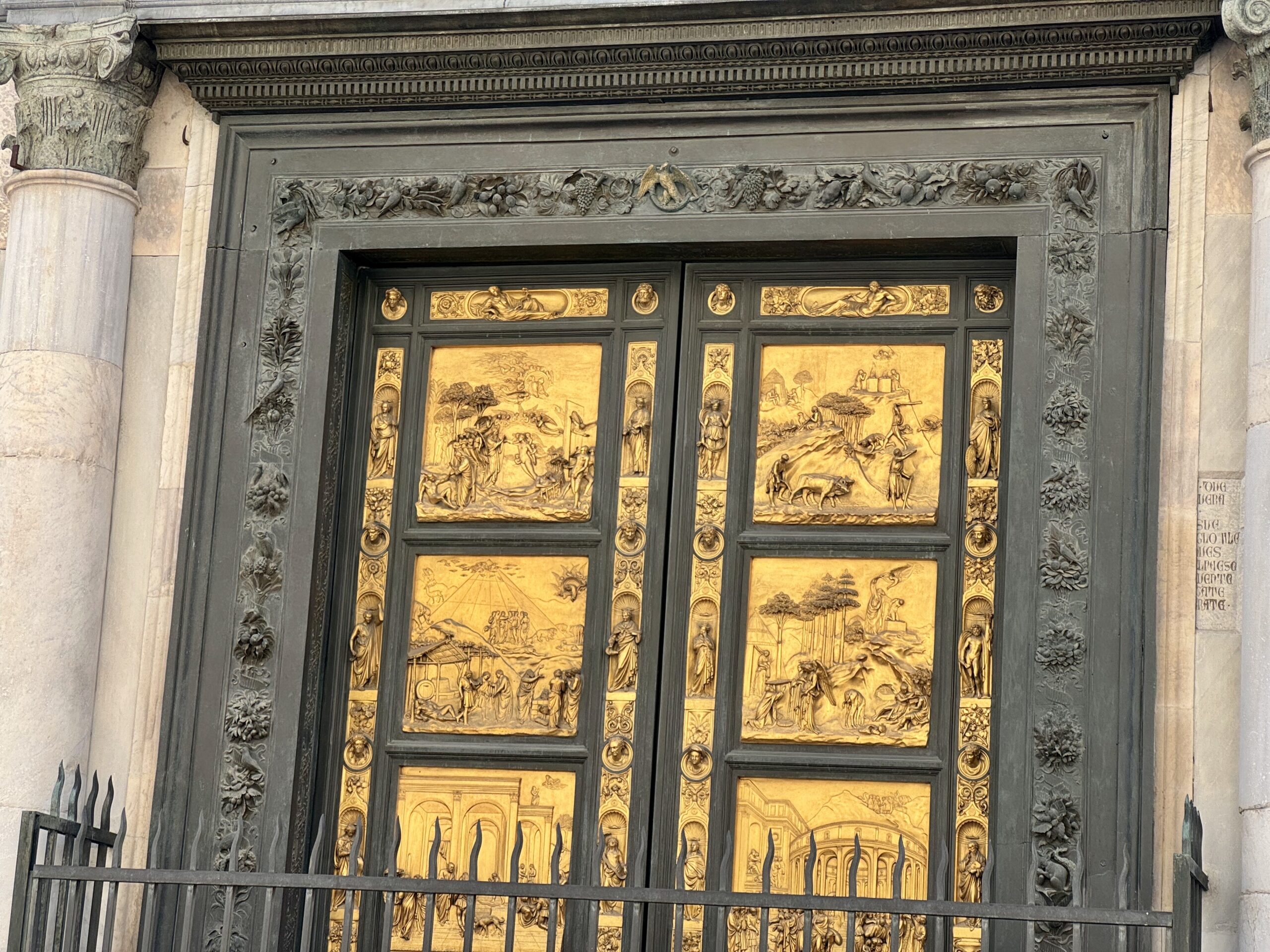Ornate metal door with gold relief panels depicting scenes, framed by carved vines and two stone columns