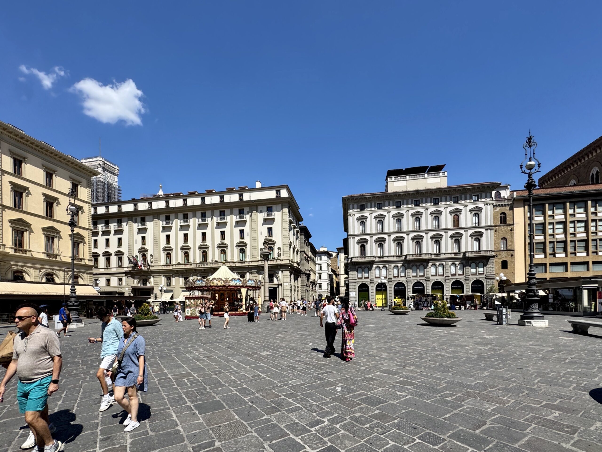 People walk across a sunlit city square with ornate historic buildings and a small carousel in the center under a blue sky.
