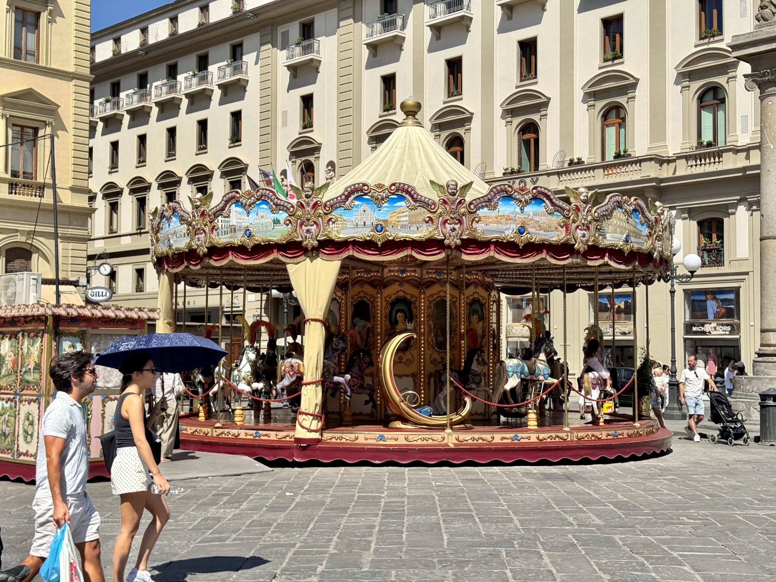 Colorful ornate carousel with horses in a city square, set against beige neoclassical buildings on a sunny day, with pedestrians nearby.