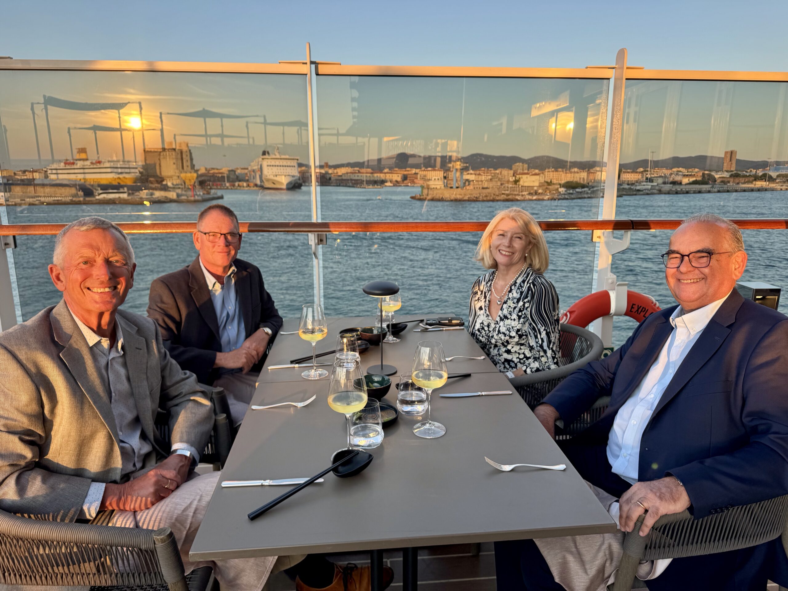 Four adults in business attire dining at a waterfront cruise, sunset over a harbor in the background, wine glasses on the table.