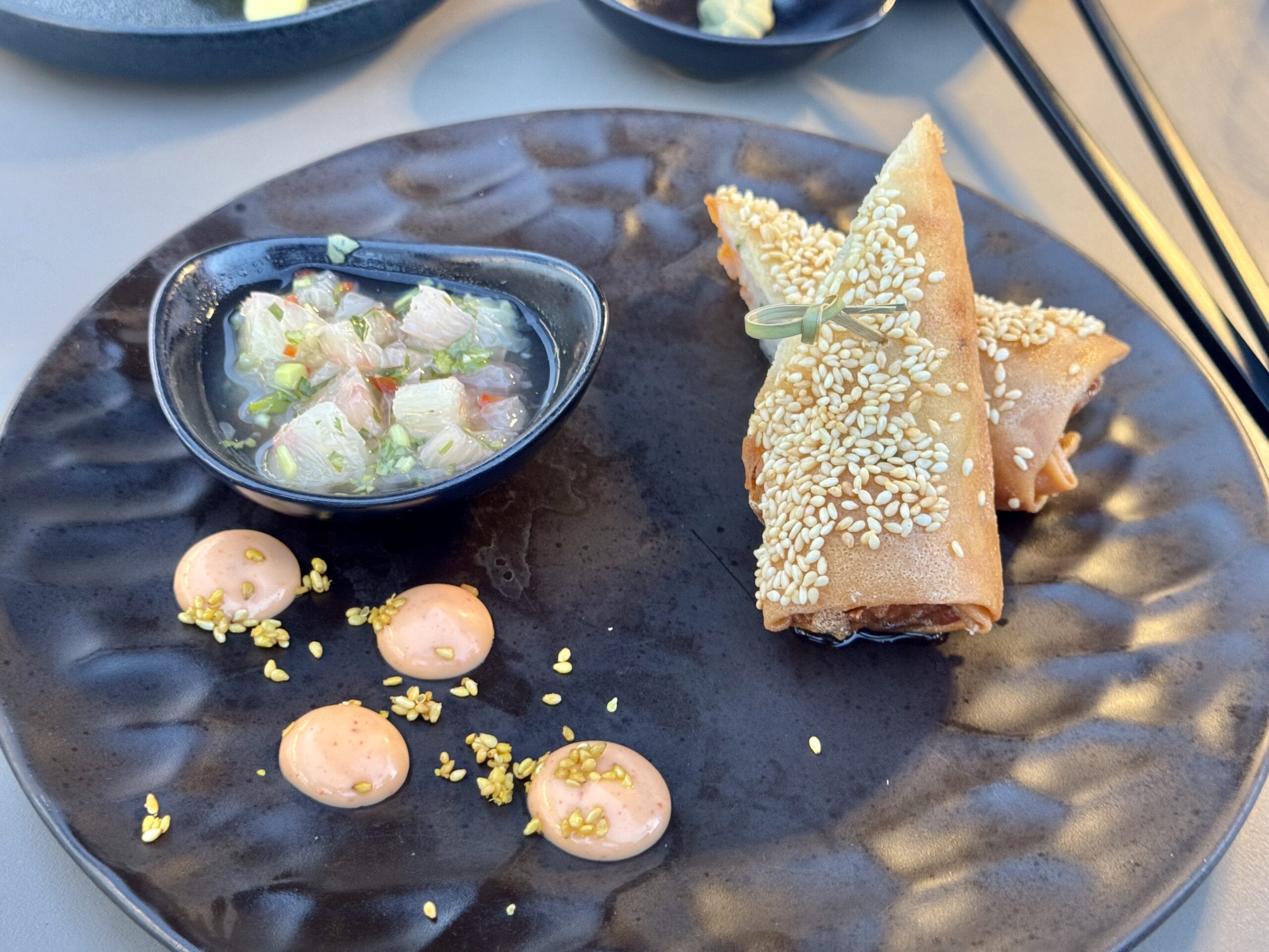 Plate with sesame-covered bread rolls and a small bowl of chopped vegetable dipping sauce, sesame seeds scattered on the plate, and pink sauce dots around the edge.