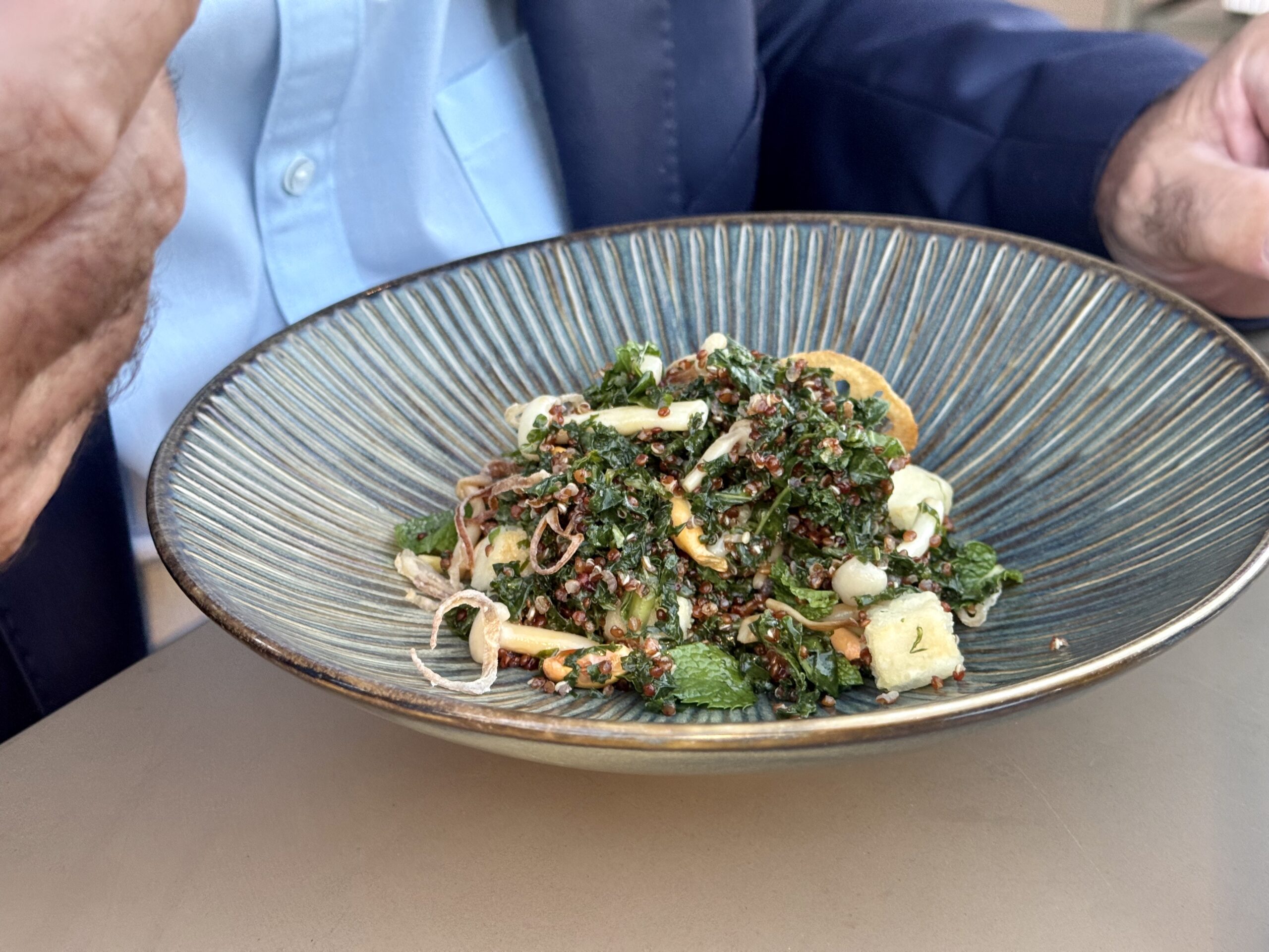 Blue ceramic plate holds a herb-packed salad with greens, quinoa, and crispy onion strands.