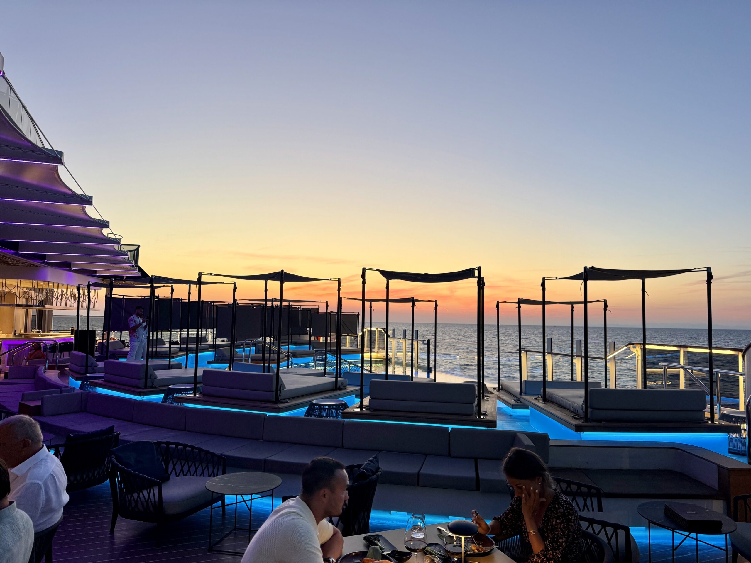 Sunset over a waterfront rooftop bar with blue-lit lounge seating and glass railings.