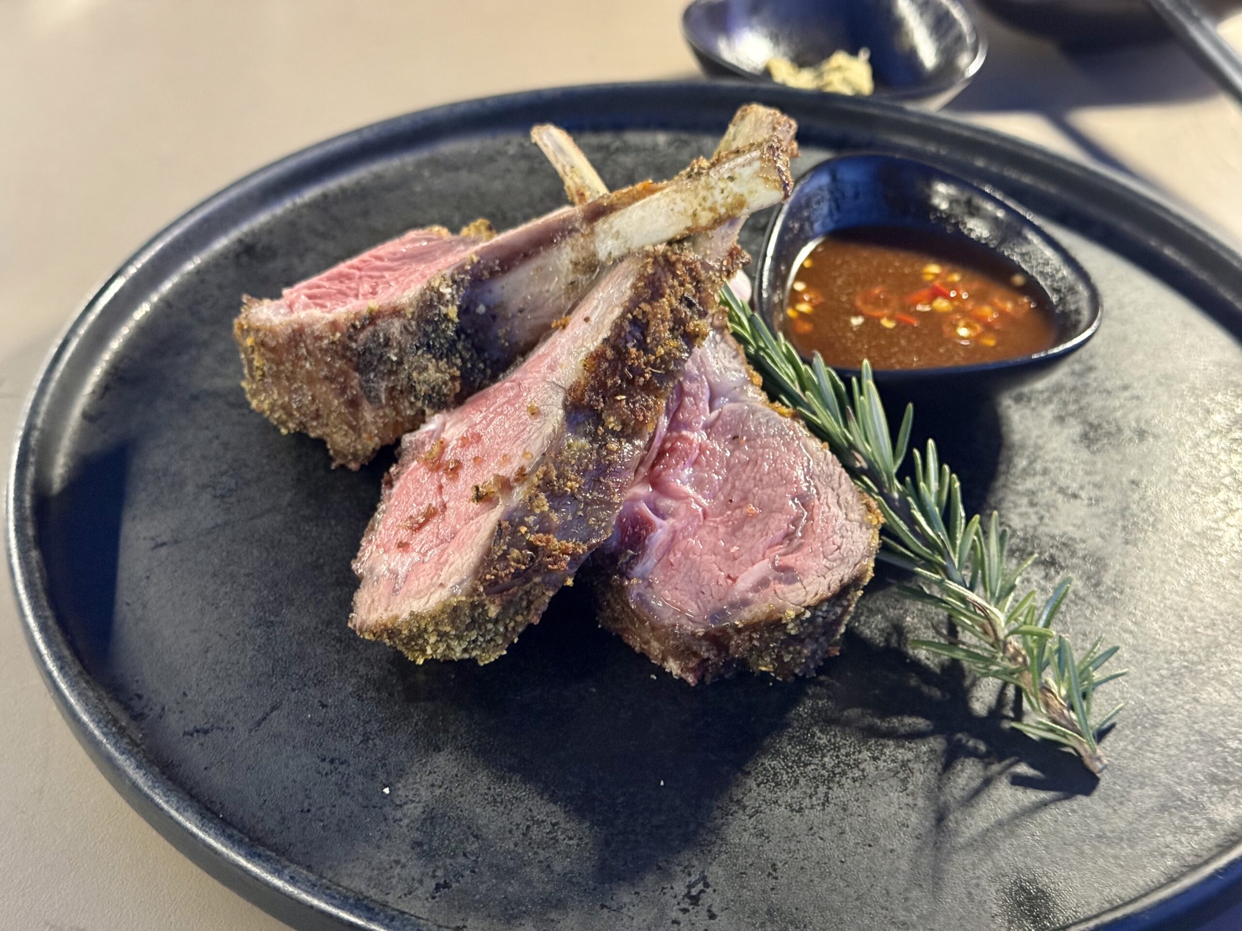 Sliced crusted ribs with pink center on a dark plate, garnished with a sprig of rosemary and a red chili sauce on the side in a small bowl
