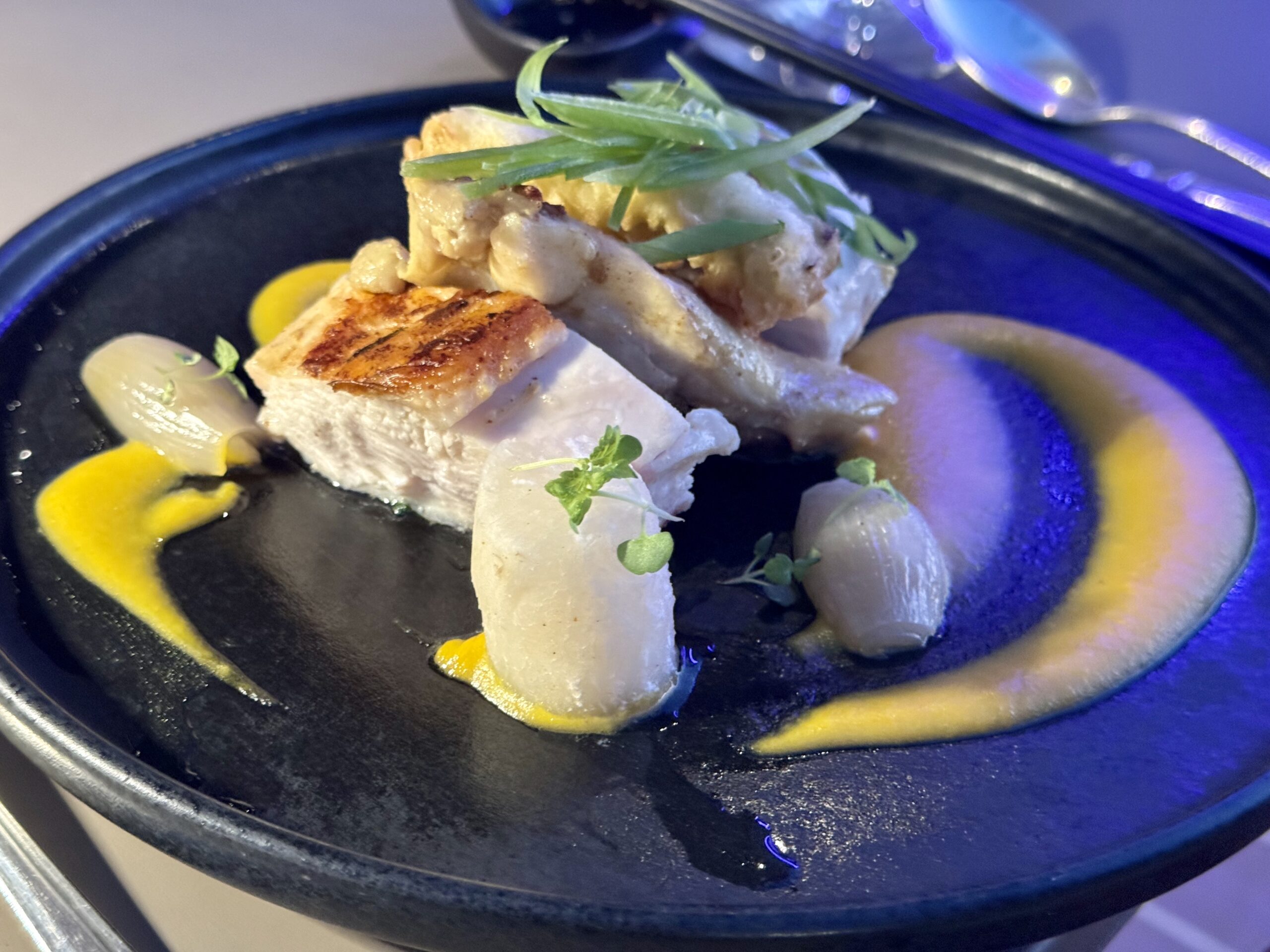 Plate with seared white fish pieces topped with green herbs and yellow sauce under blue lighting.