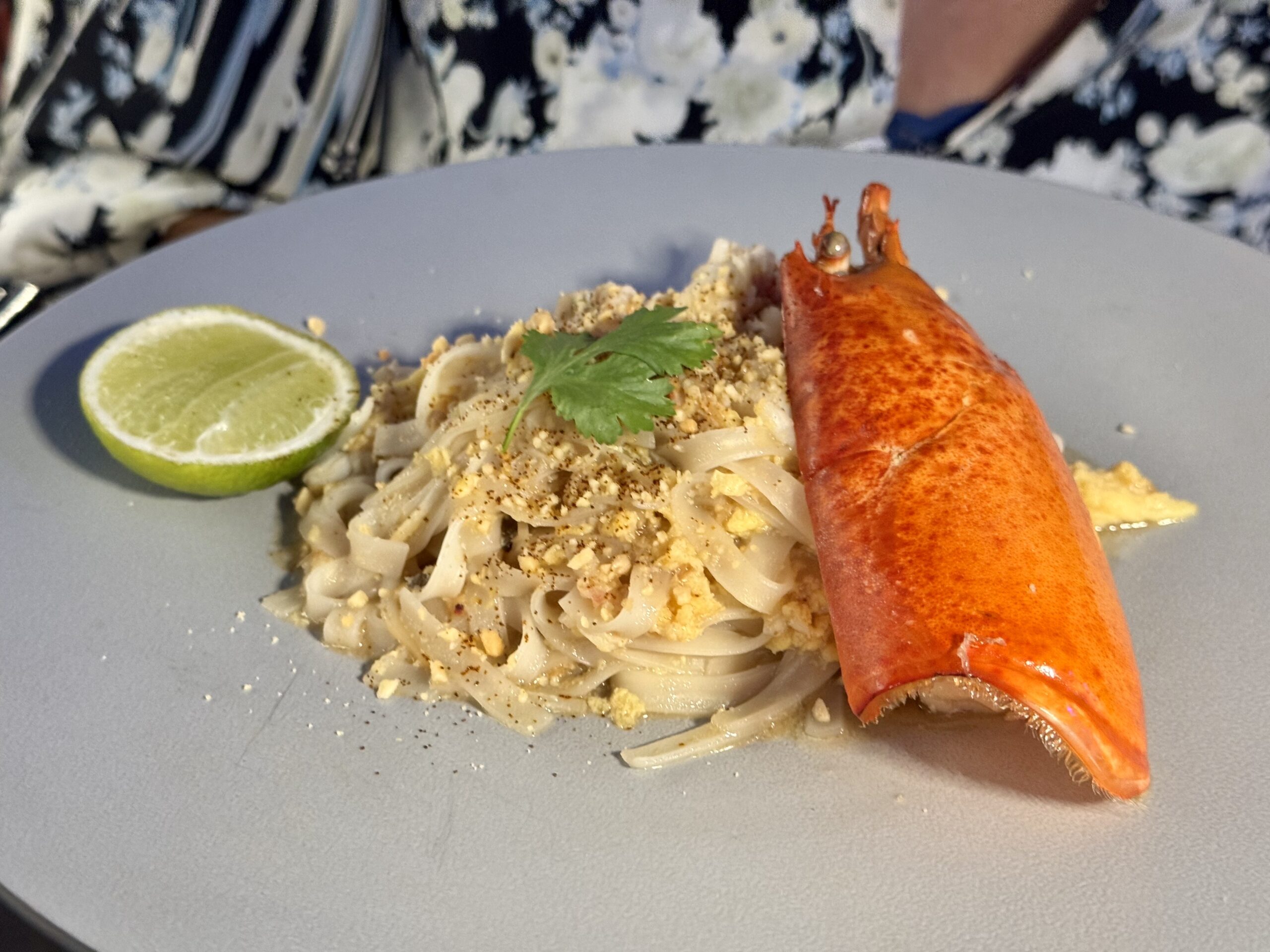 Lobster tail rests on a plate beside pasta with lime wedge and cilantro garnish.