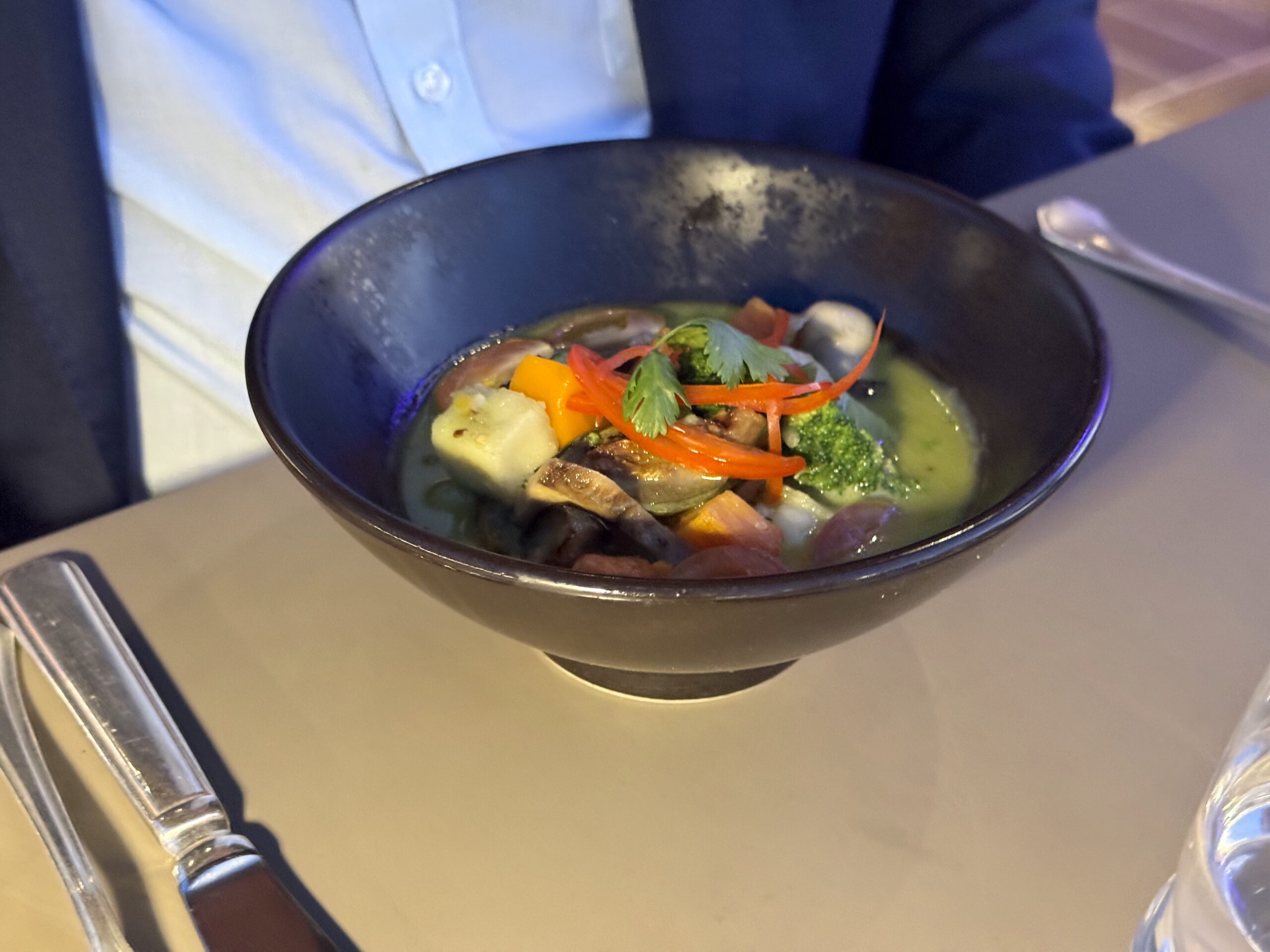 Dark bowl of vegetable soup with carrot ribbons, broccoli, potato chunks, and cilantro on a table, with cutlery nearby.