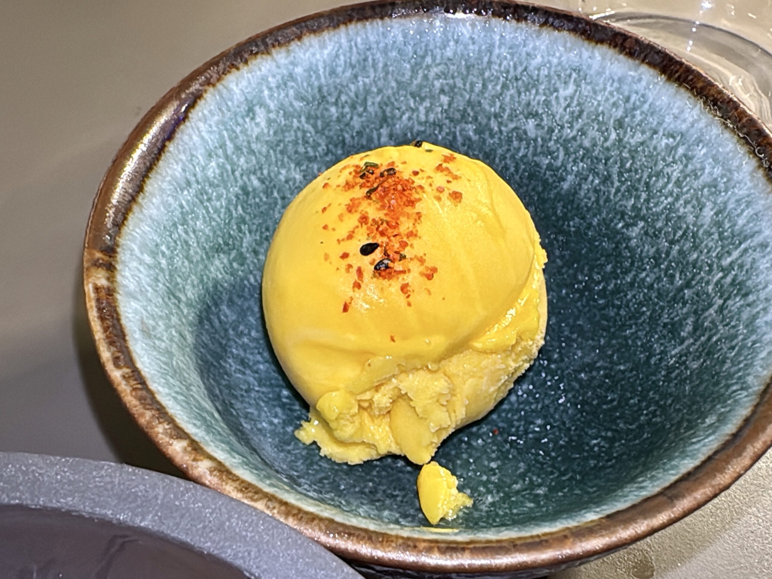 Close-up of a yellow pastry or scoop in a blue ceramic bowl with orange-red specks on top.