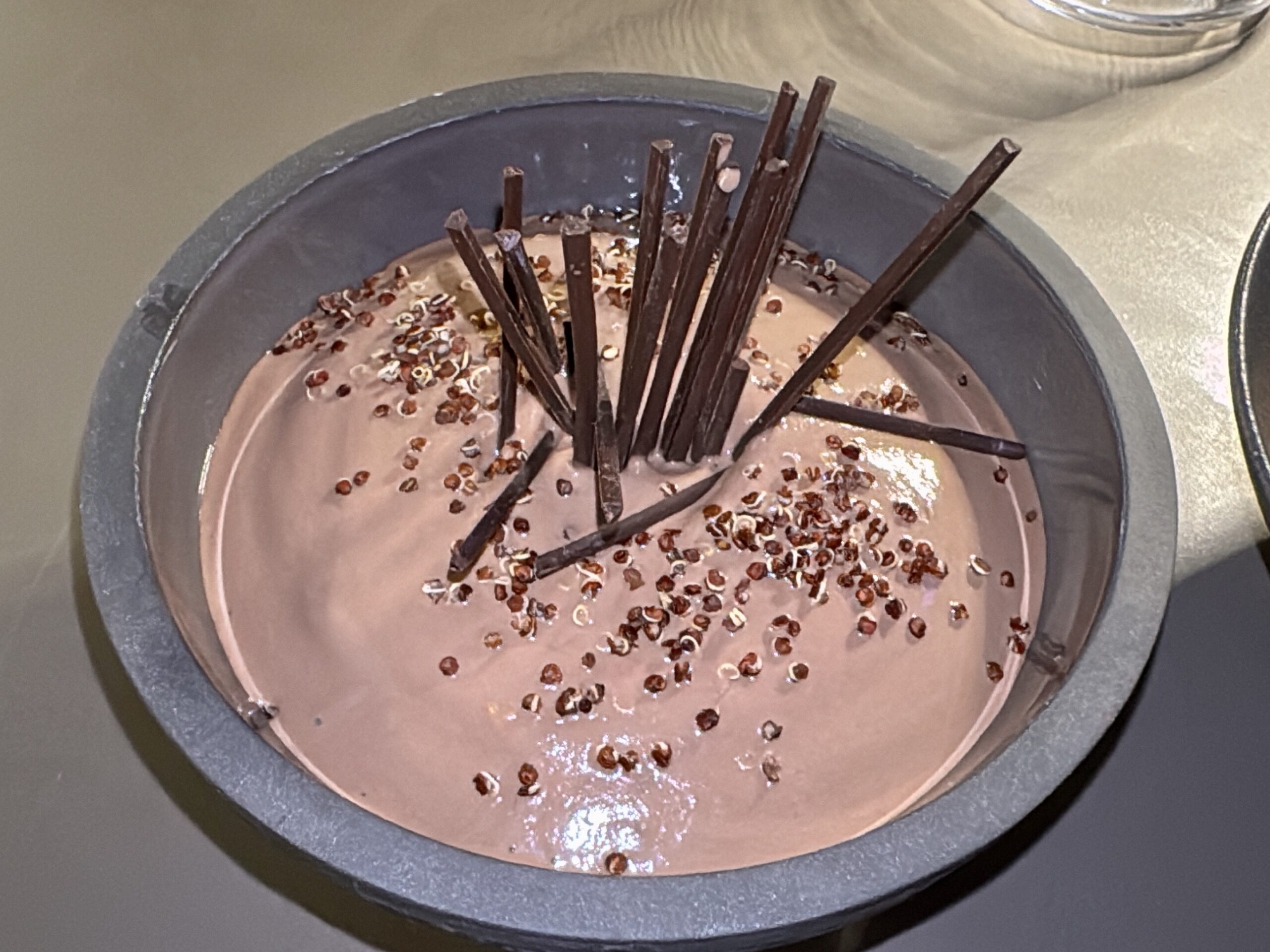 Round chocolate cake in a pan topped with upright chocolate sticks and sprinkled seeds on glossy frosting