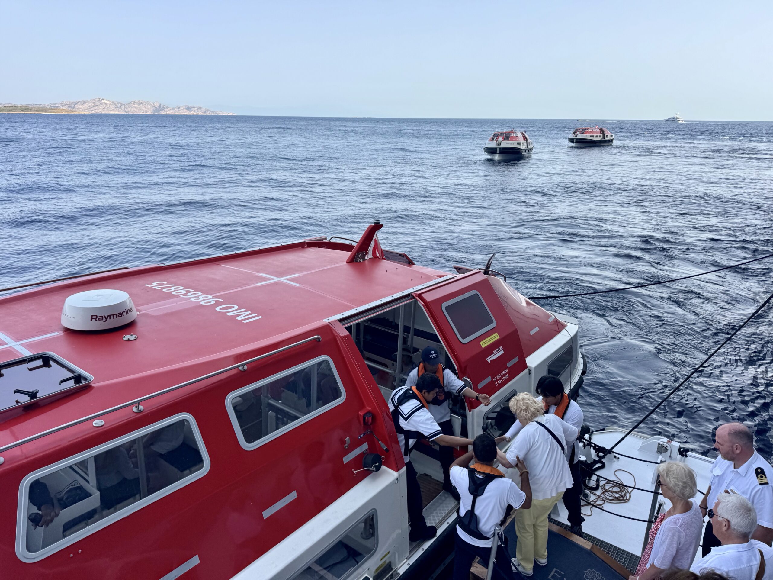 Red rescue boat at sea with passengers wearing life jackets boarding the vessel; calm blue water and distant boats on the horizon.
