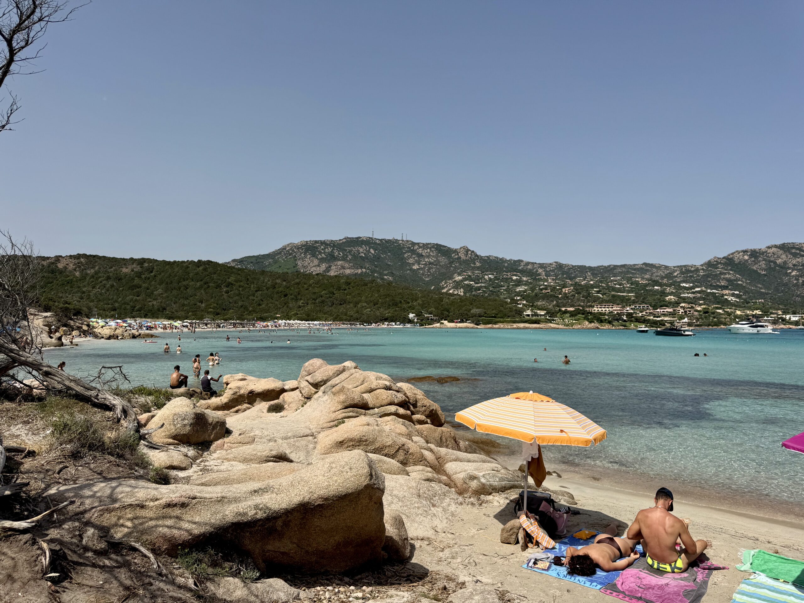People sunbathing on a rocky beach with turquoise water and green hills in the background, under a clear blue sky.