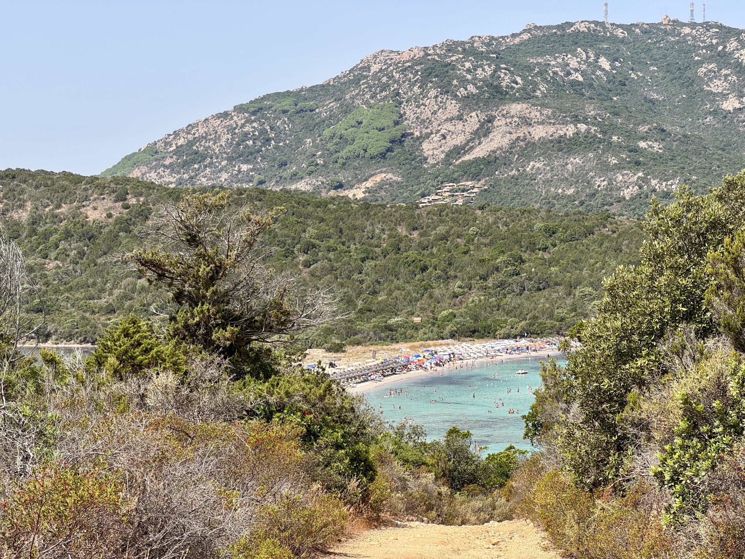Coastal beach scene with turquoise water, sunbathers along the shore, and green hills in the background.