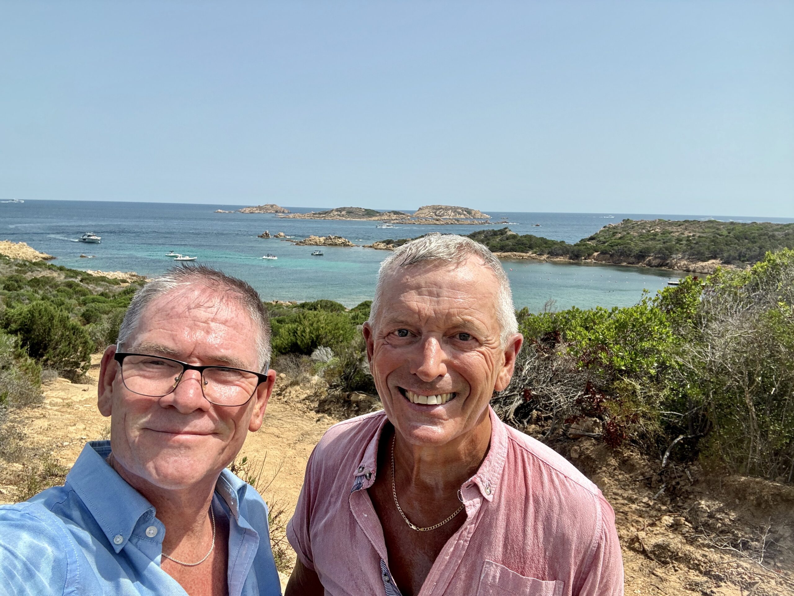 Two men taking a seaside selfie with rocky coastline, blue sky, and turquoise water behind them. The man on the left wears a light blue shirt and glasses; the man on the right wears a pink shirt and necklace.