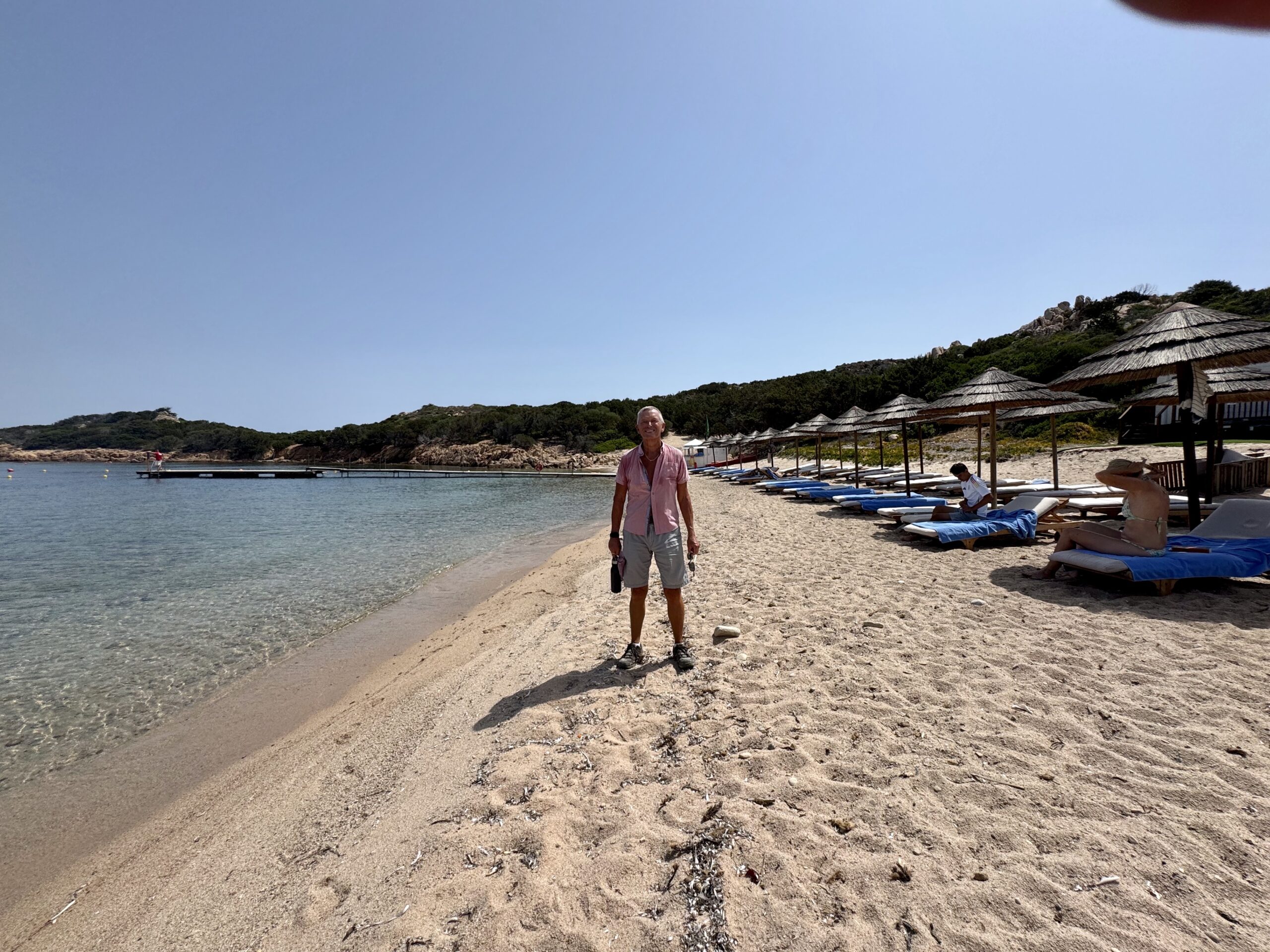 Man stands on a sandy beach near calm blue water, with thatched umbrellas and lounge chairs along the shore behind him.