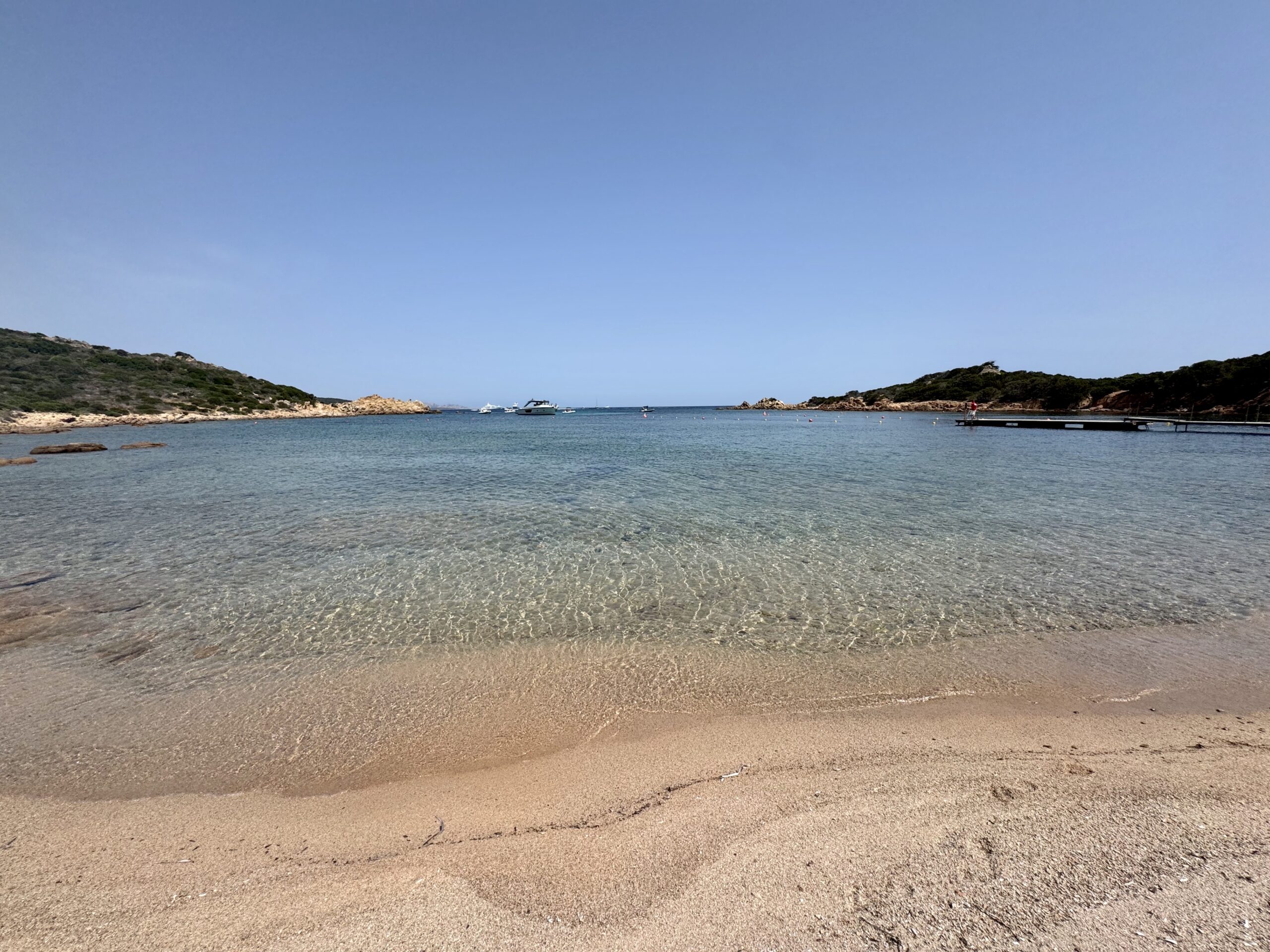 Bright blue sky over a calm bay, with rocky, green hills along the shore and several small boats floating near the horizon; a dock extends from the right side into the water along the shore.