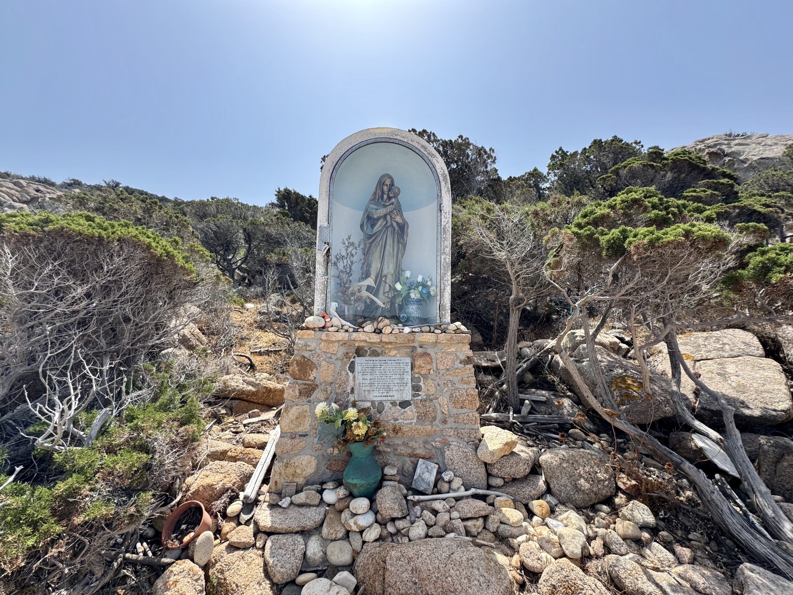 Statue of Mary in a rounded glass shrine set on a stone pedestal among rocks and scrubby bushes under a clear blue sky.